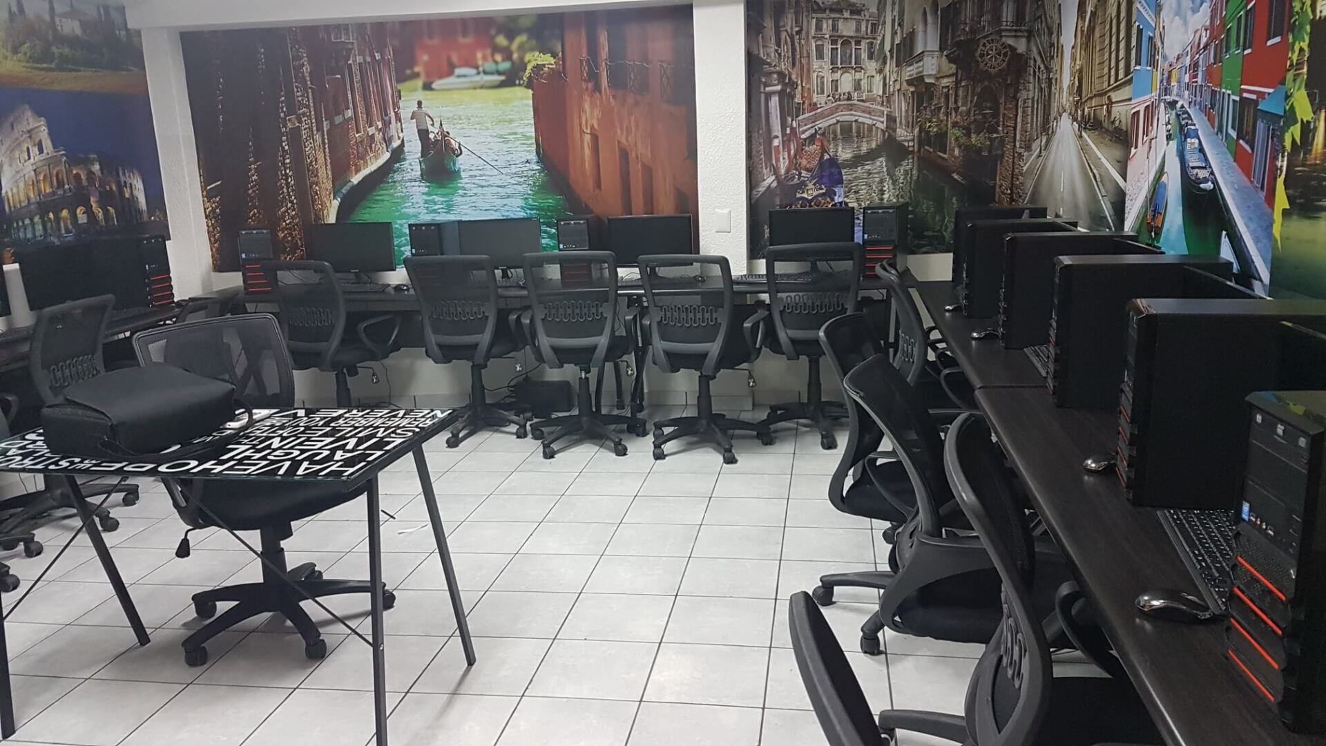 Sala de informática vacía con filas de sillas y mesas negras, suelo de baldosas y carteles en la pared al fondo.