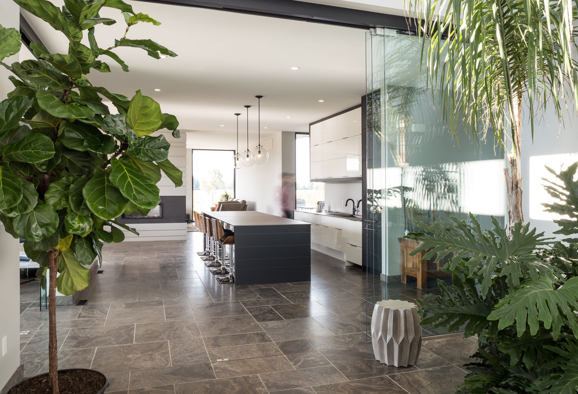 Open-concept home with kitchen island, indoor plants, and a glass-walled space. Dark tile floors.