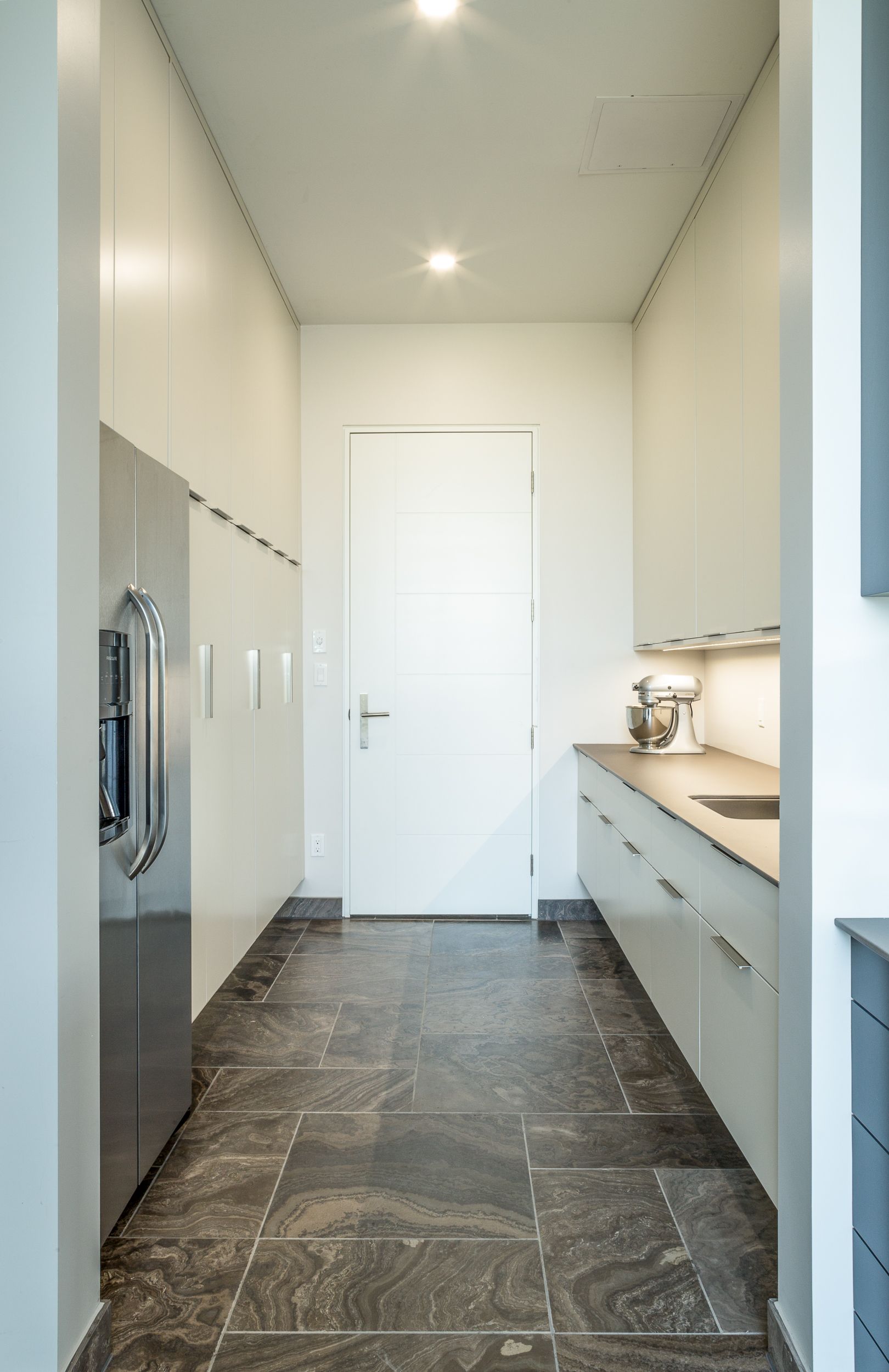 Narrow kitchen with dark stone floor, white cabinets, stainless steel fridge, and a closed white door.