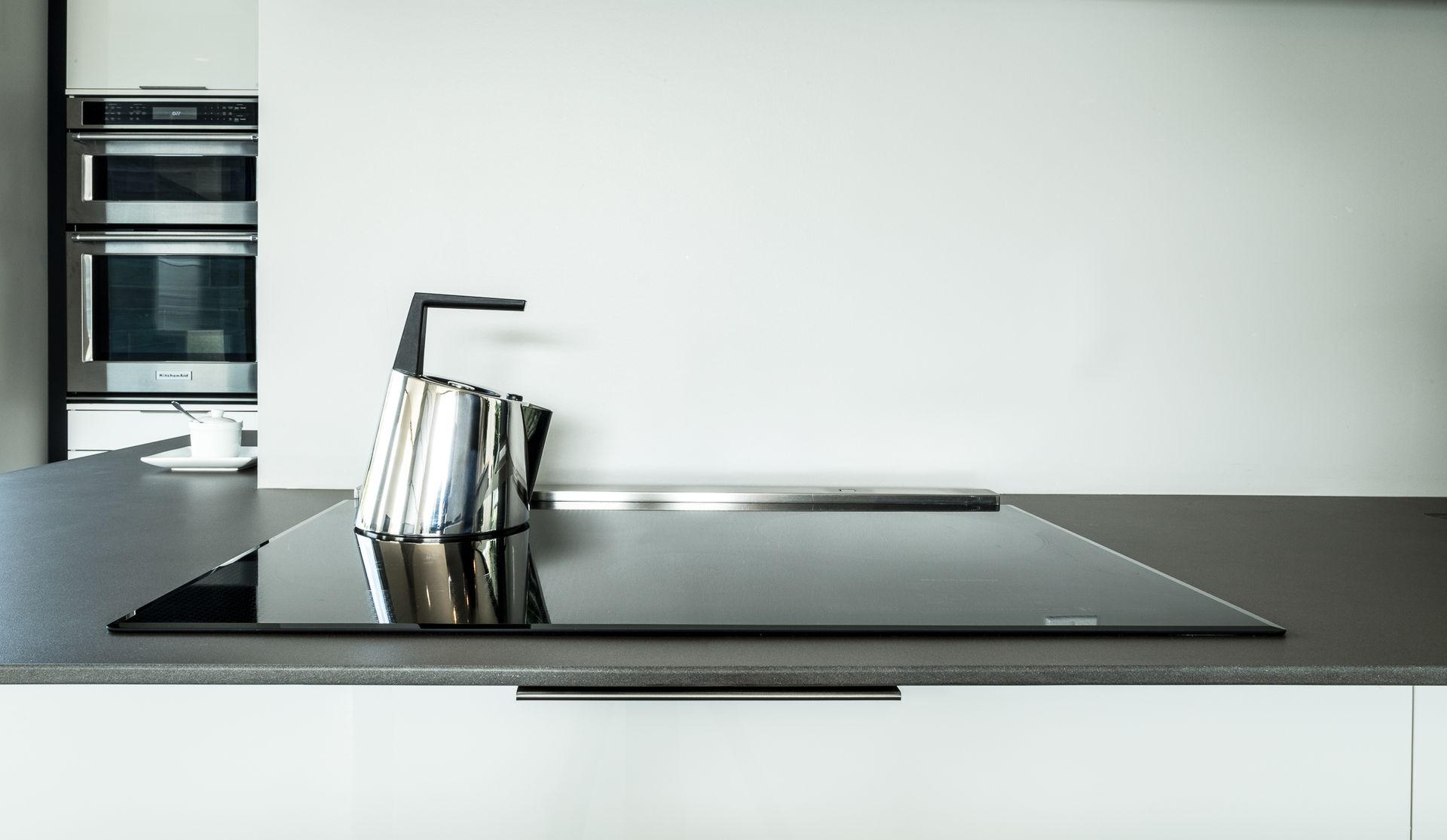 Stainless steel tea kettle on a black cooktop in a modern kitchen with built-in ovens.