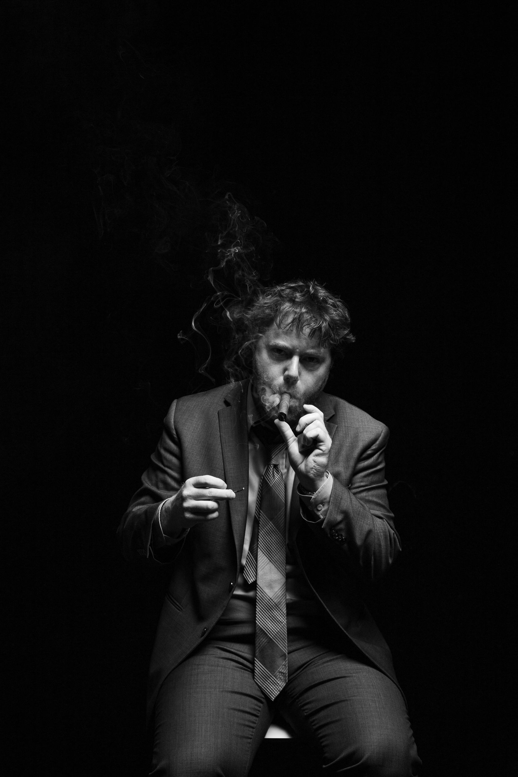 Man in suit, seated, smoking cigar, smoke rising, dark background.
