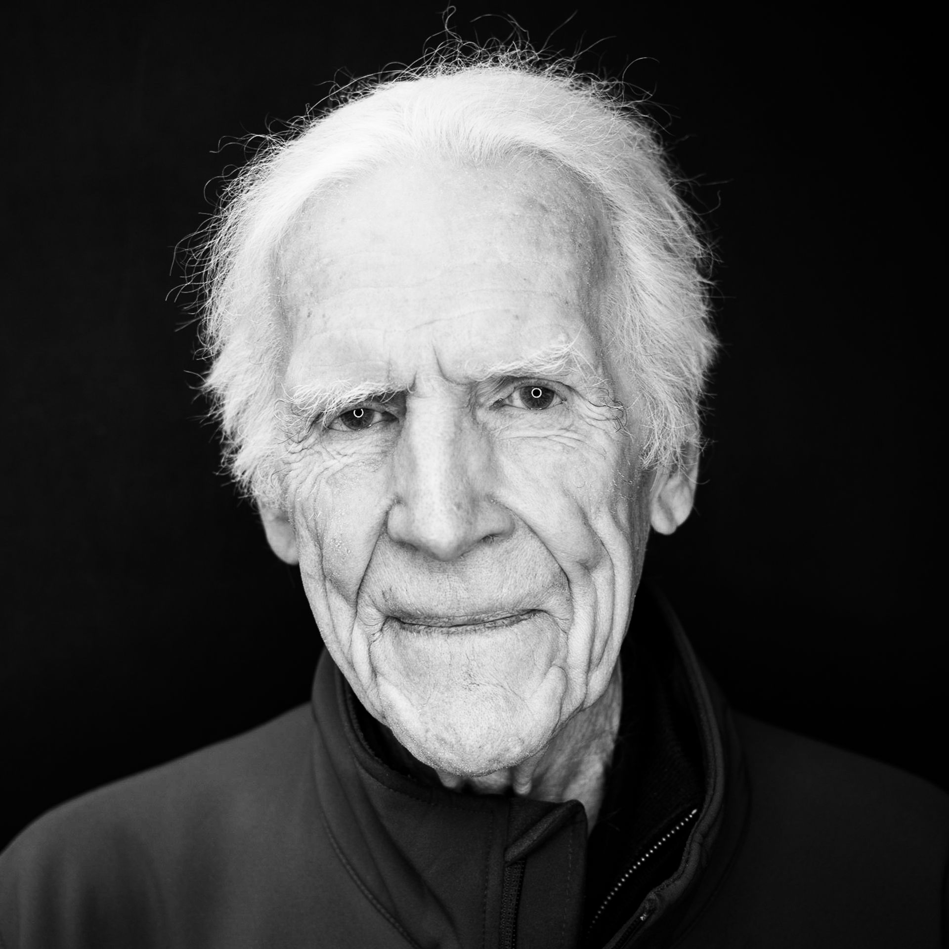 Senior man with white hair, smiling, in front of a dark backdrop.