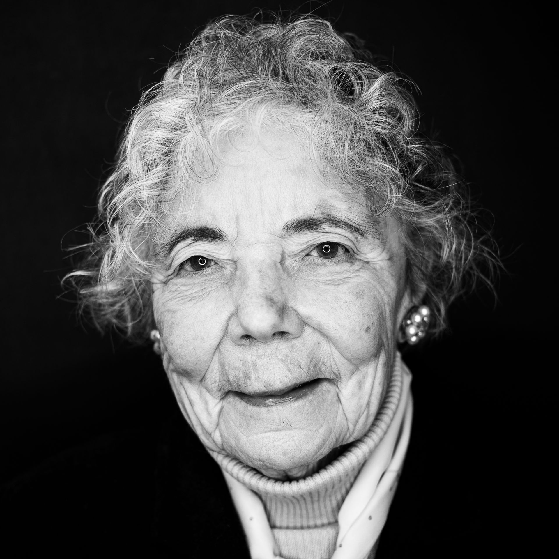 Elderly woman with curly white hair, smiling. Wearing a turtleneck and pearl earrings, black background.
