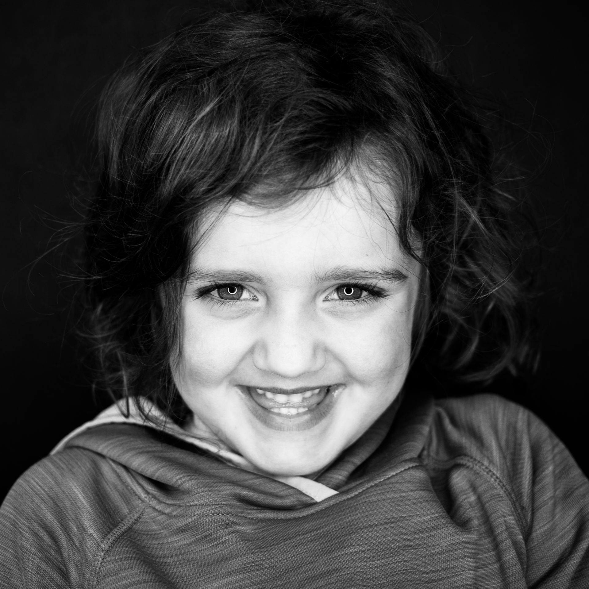 Smiling child with curly hair wearing a hoodie against a black backdrop.