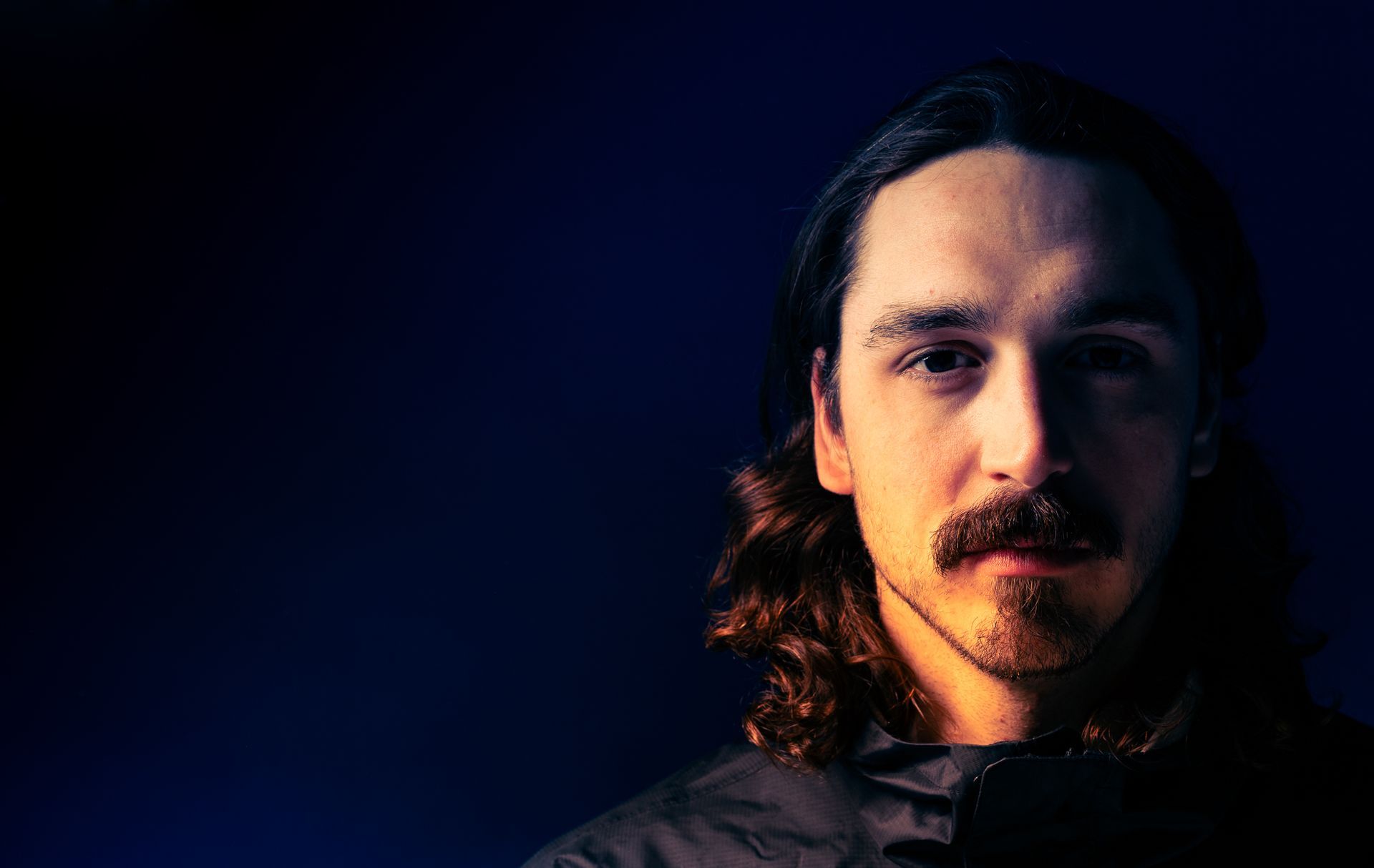 Man with a mustache and long hair, lit from the side, with a blue background.