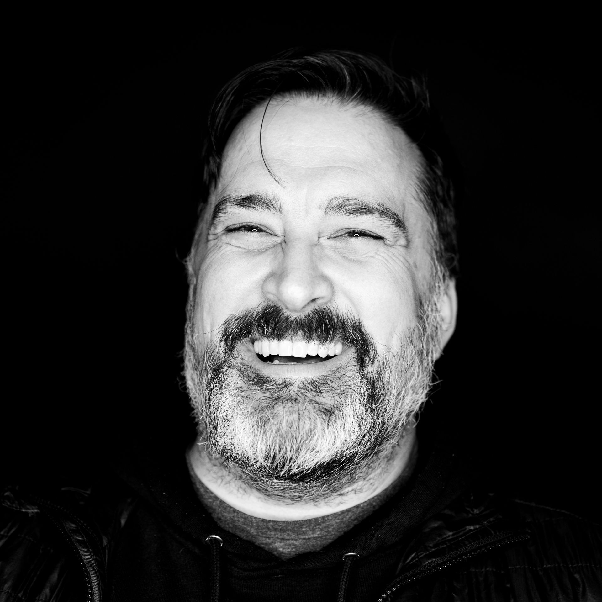 Man with a beard smiling, eyes closed, against a black background.