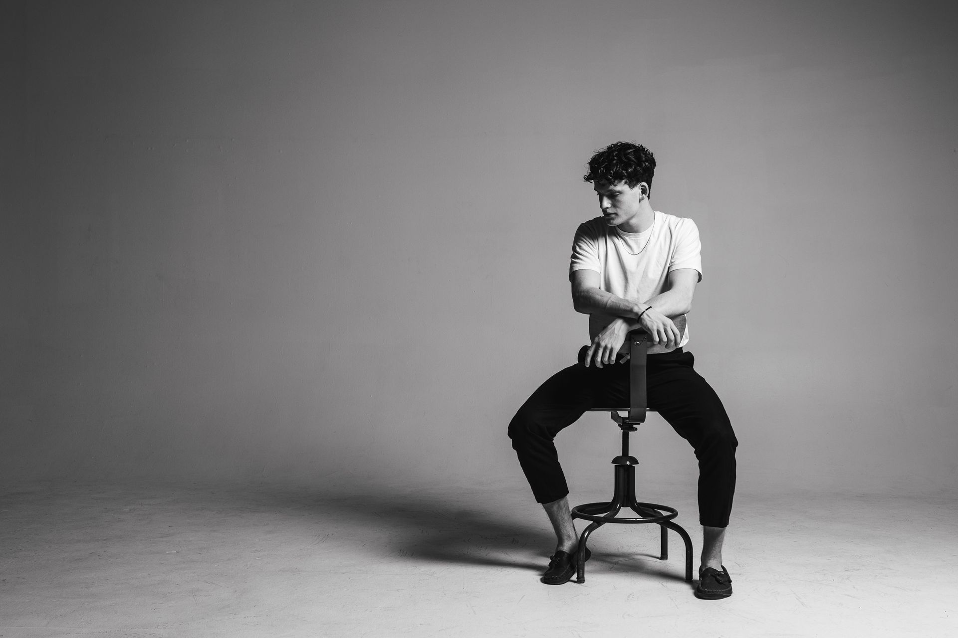 Man in casual attire sitting on a stool, looking down. Black and white, studio background.