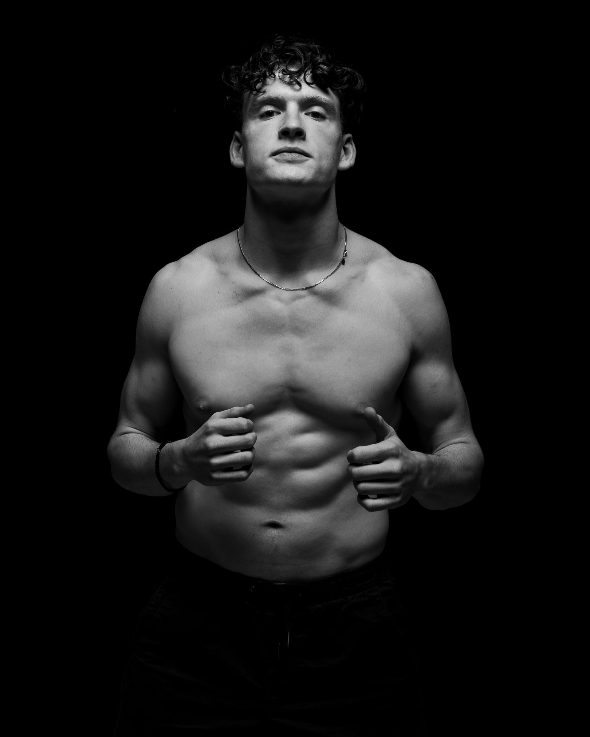 Muscular person with curly hair, shirtless, holding hands near torso, in shadow.
