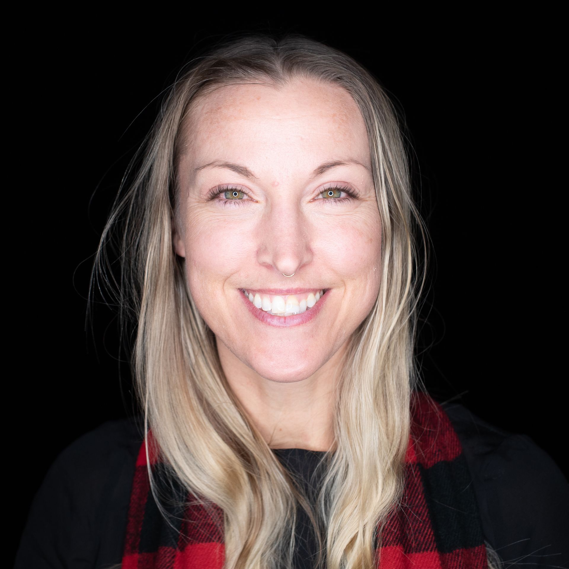 Woman with long blonde hair smiles, wearing a black shirt and red plaid scarf.