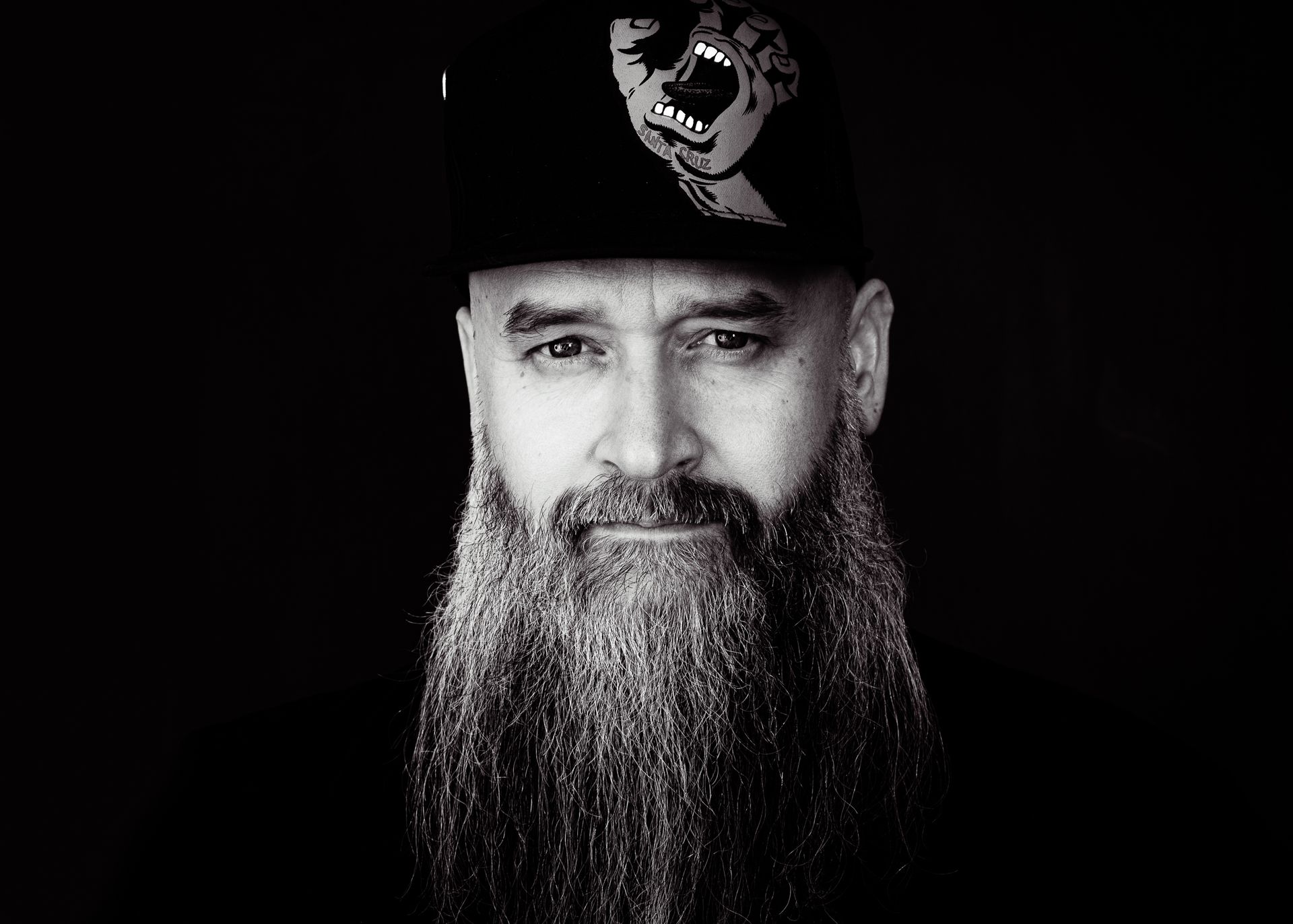 Man with long gray beard and black hat with dog design, staring at the viewer.