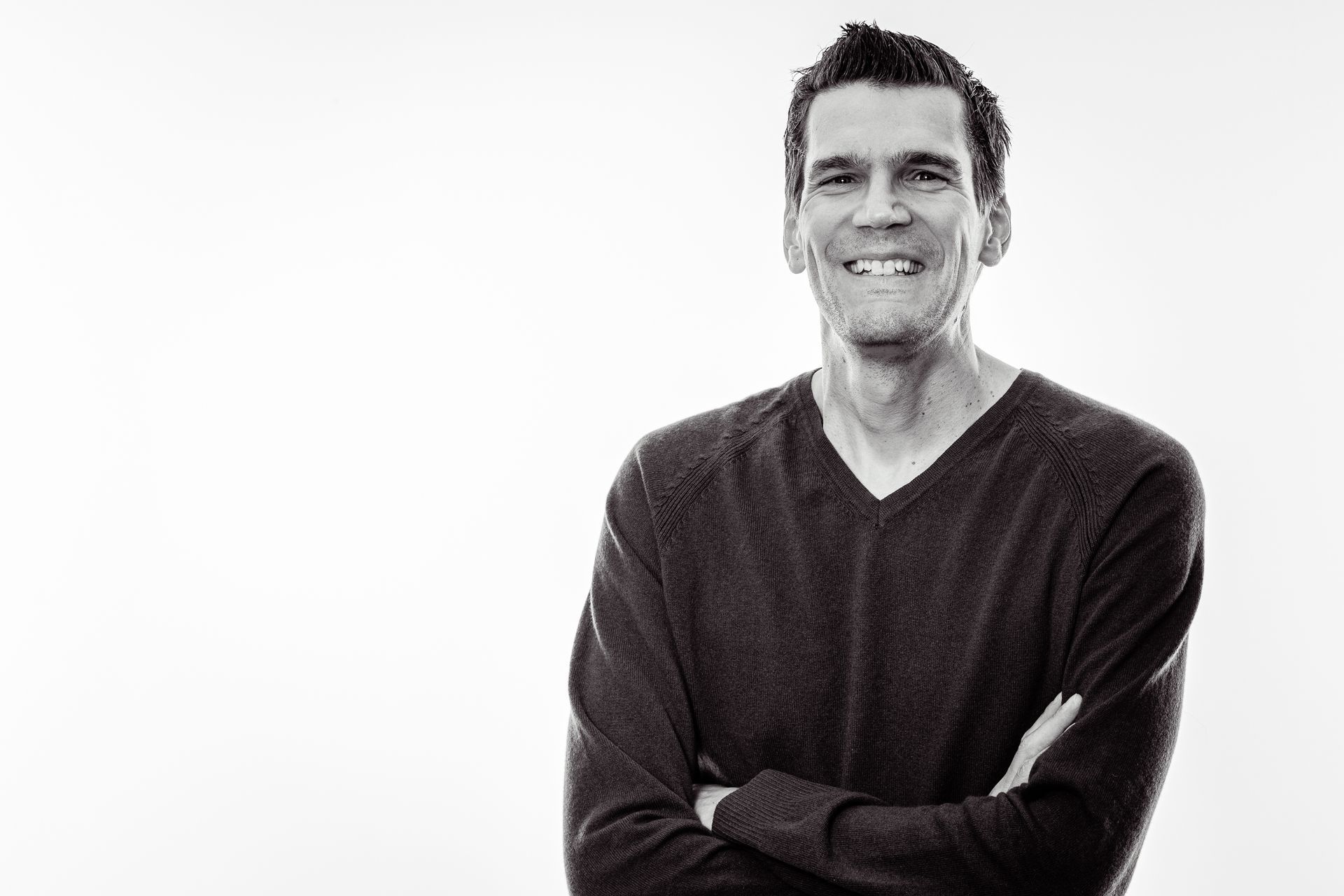Smiling man with arms crossed, wearing a dark v-neck sweater, against a white background.