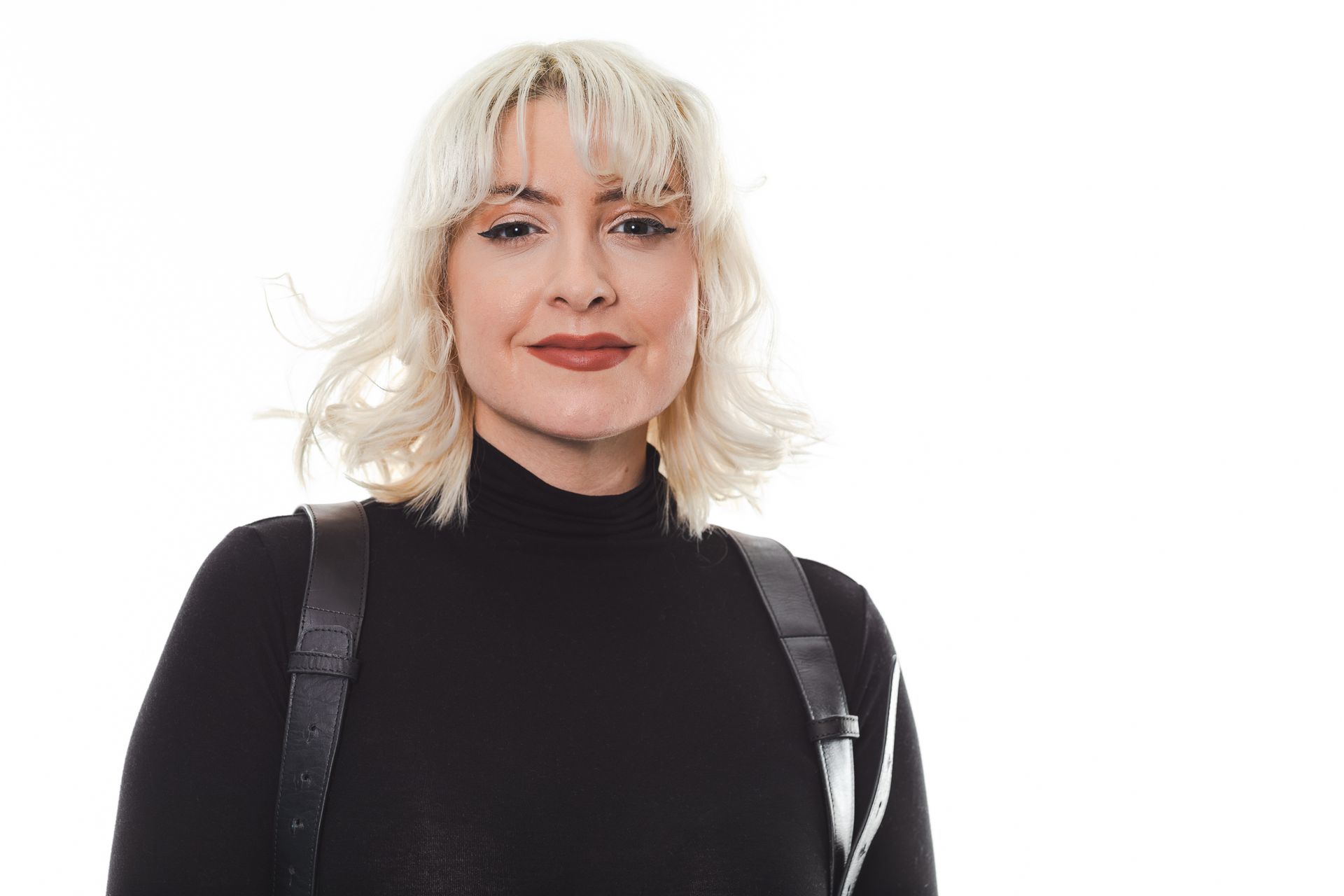 Woman with blonde hair wearing a black turtleneck and leather straps, smiling against a white background.