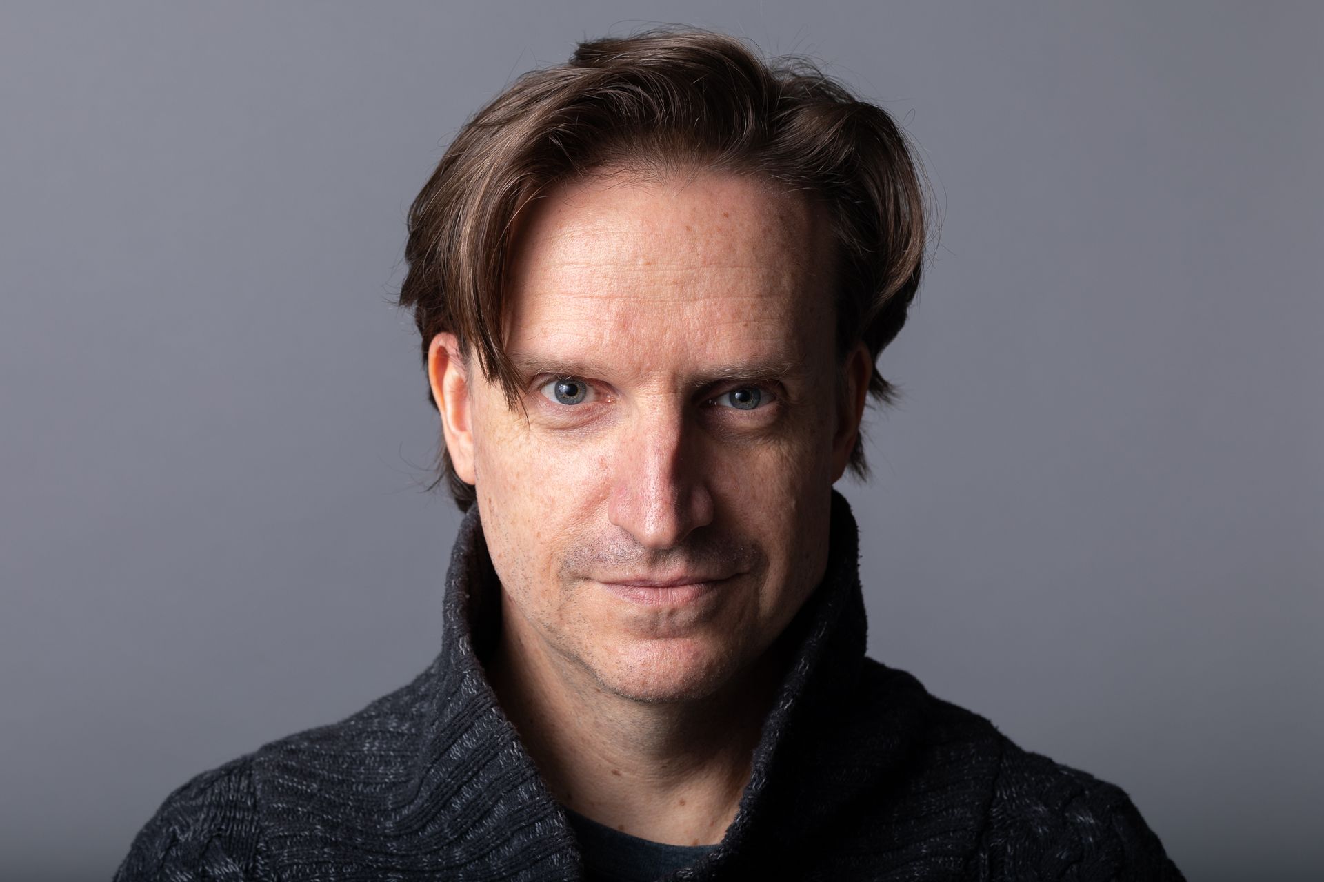 Man with light skin and brown hair looking at the camera, wearing a dark sweater against a gray background.
