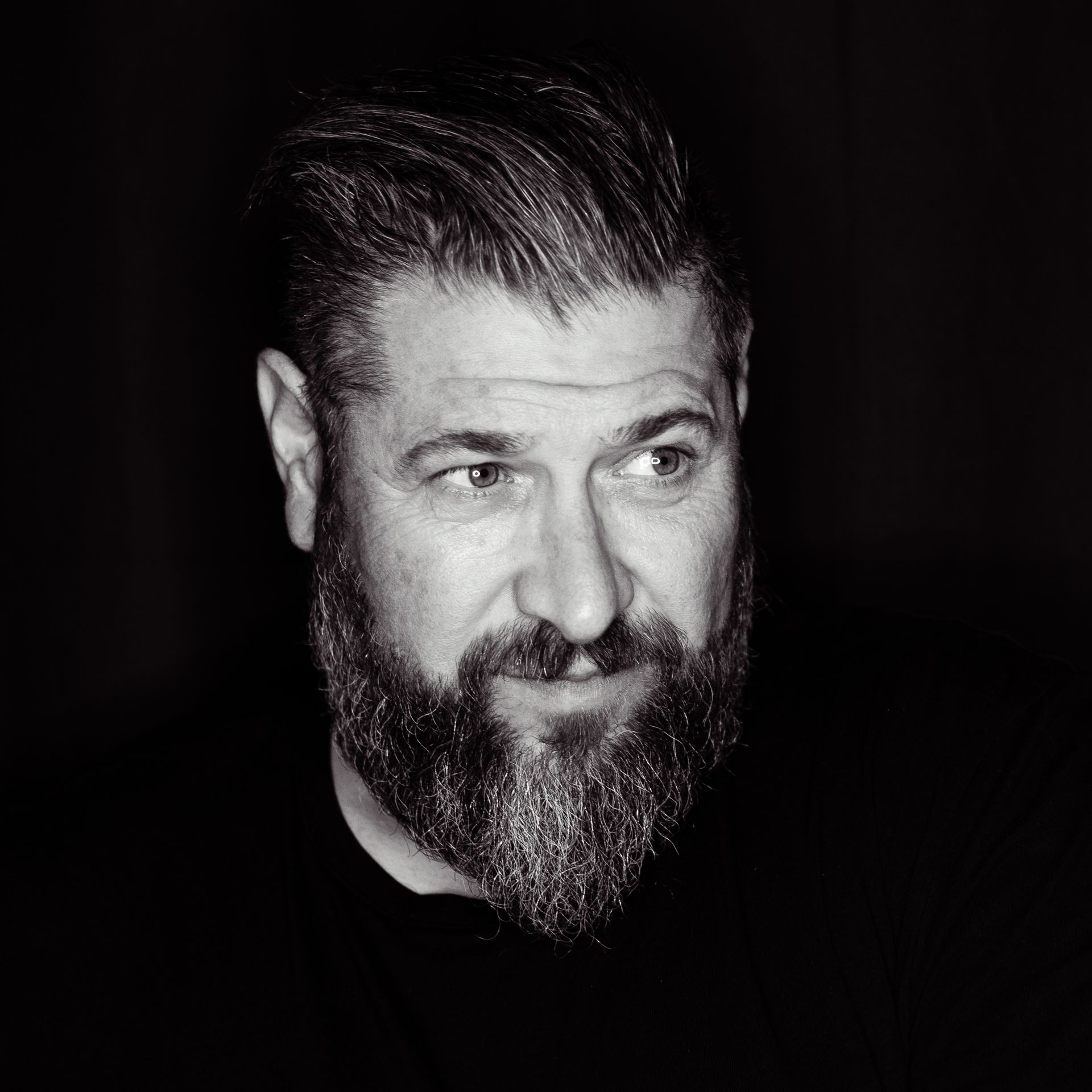 Man with full beard, swept-back hair, looking to the side, wearing a black shirt. Black and white photo.