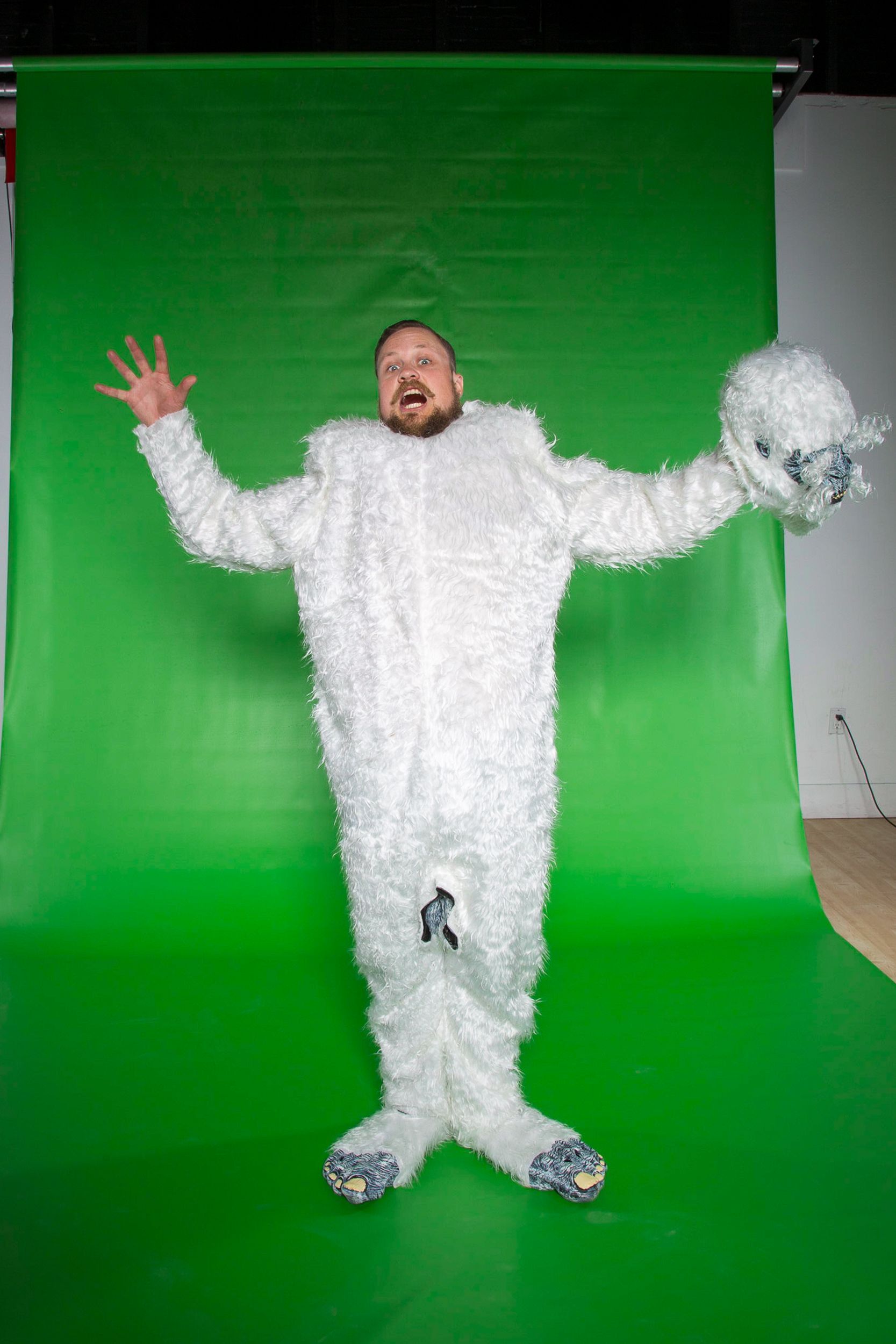 Man in white, furry costume and holding a head, stands against a green backdrop with arms outstretched.