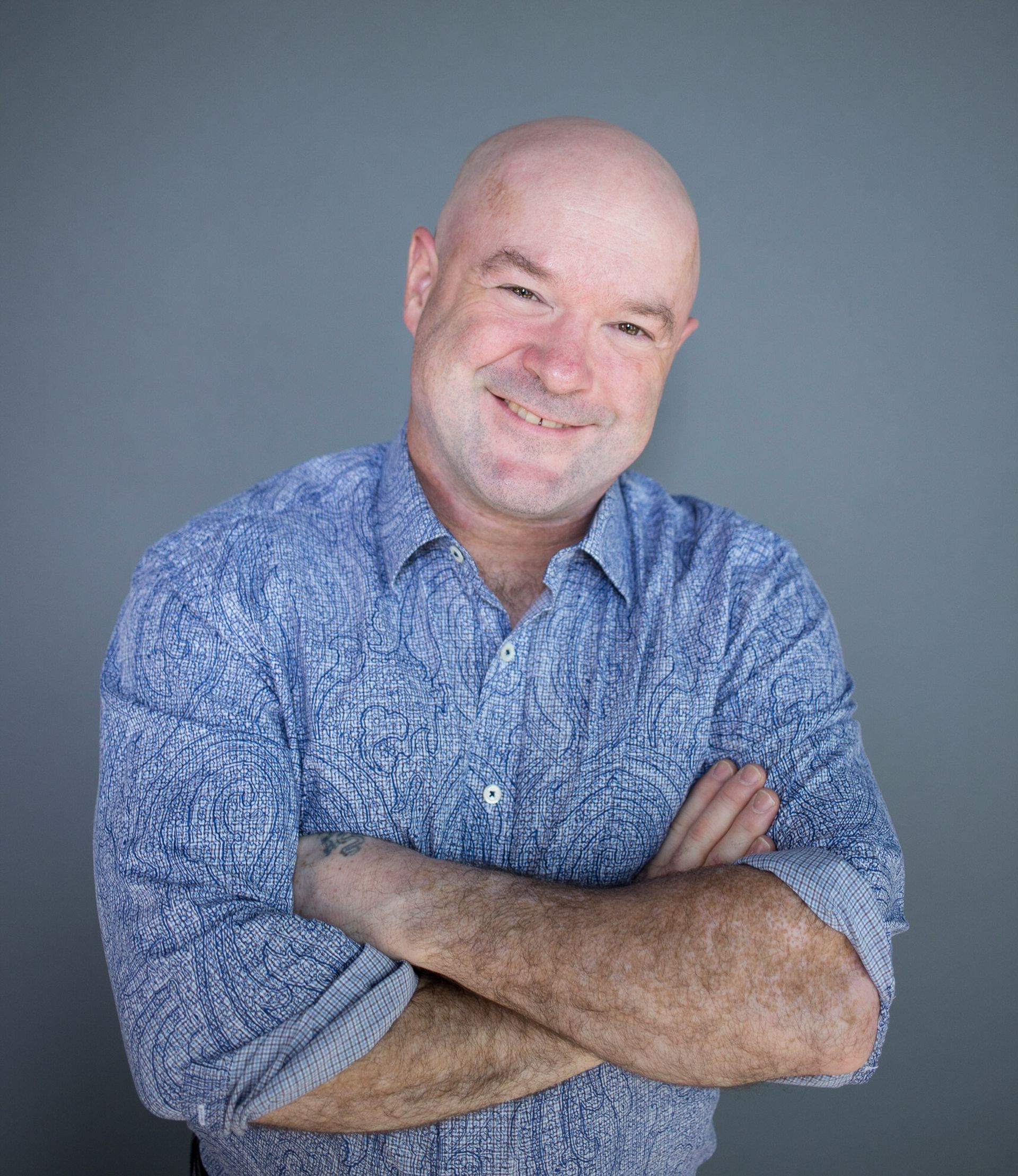 Bald man in blue patterned shirt smiles, arms crossed against gray backdrop.