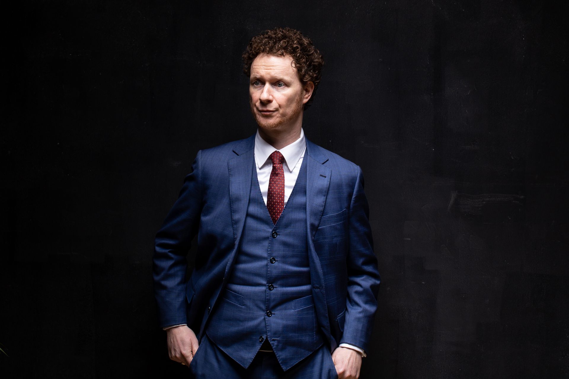 Man in blue suit, red tie, hands in pockets, looking to the side, dark background.