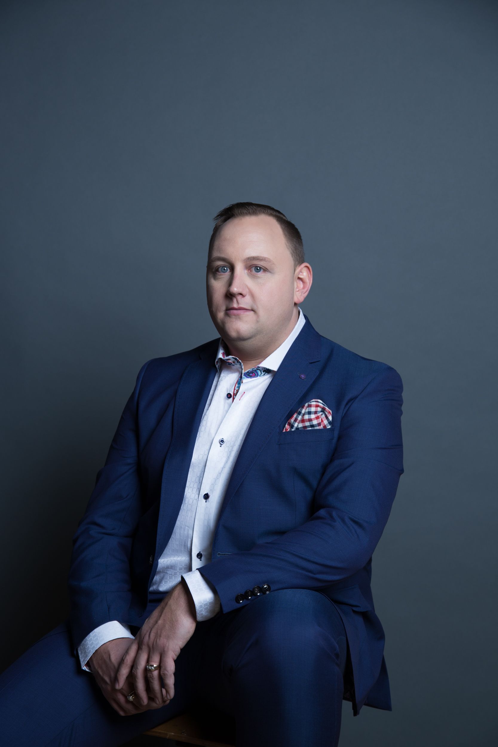 Man in blue suit, white shirt, and patterned pocket square, seated with gray background.