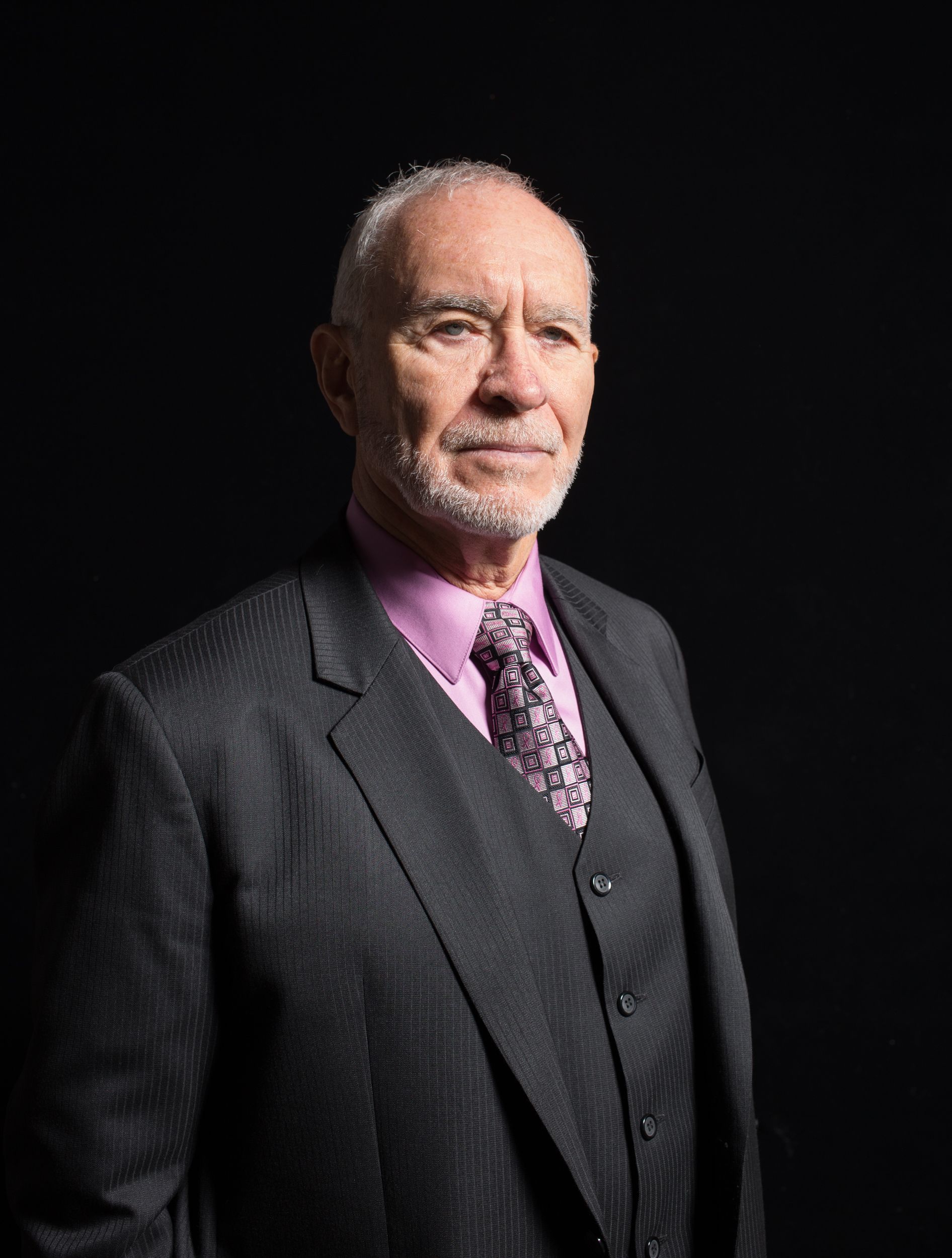Man in suit, with gray hair and beard, wearing a pink shirt and patterned tie, against a dark background.