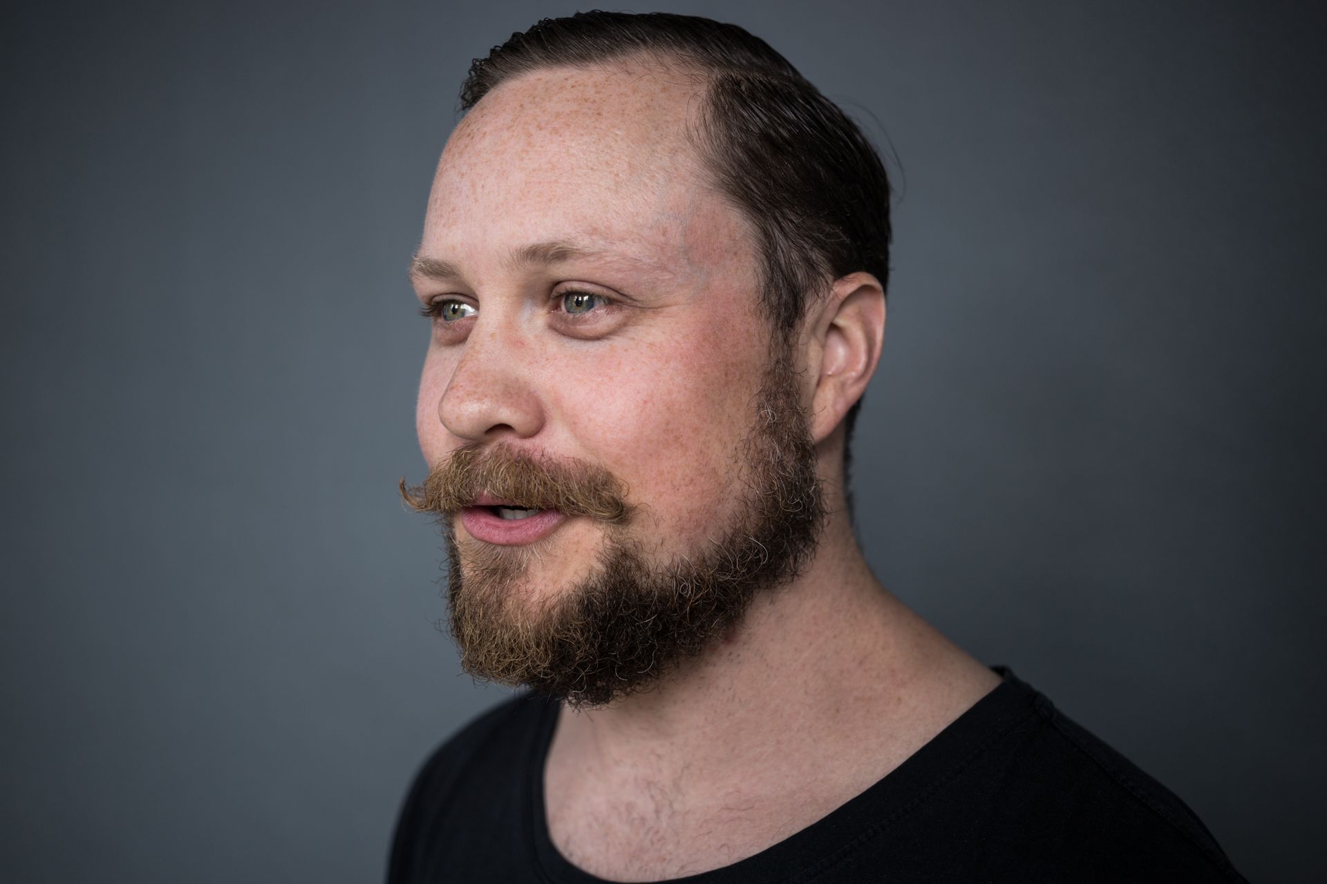 Man with brown hair and beard, looking left. He wears a black shirt, against a gray background.