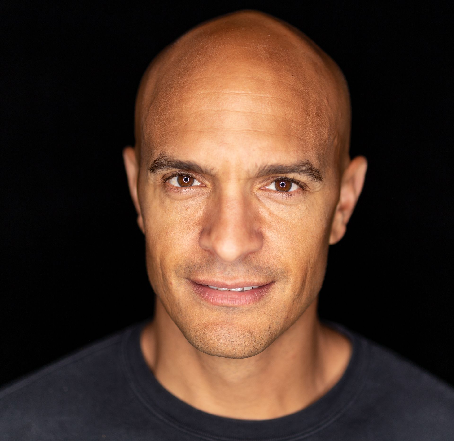 Bald man with brown eyes smiles at the camera against a dark background.