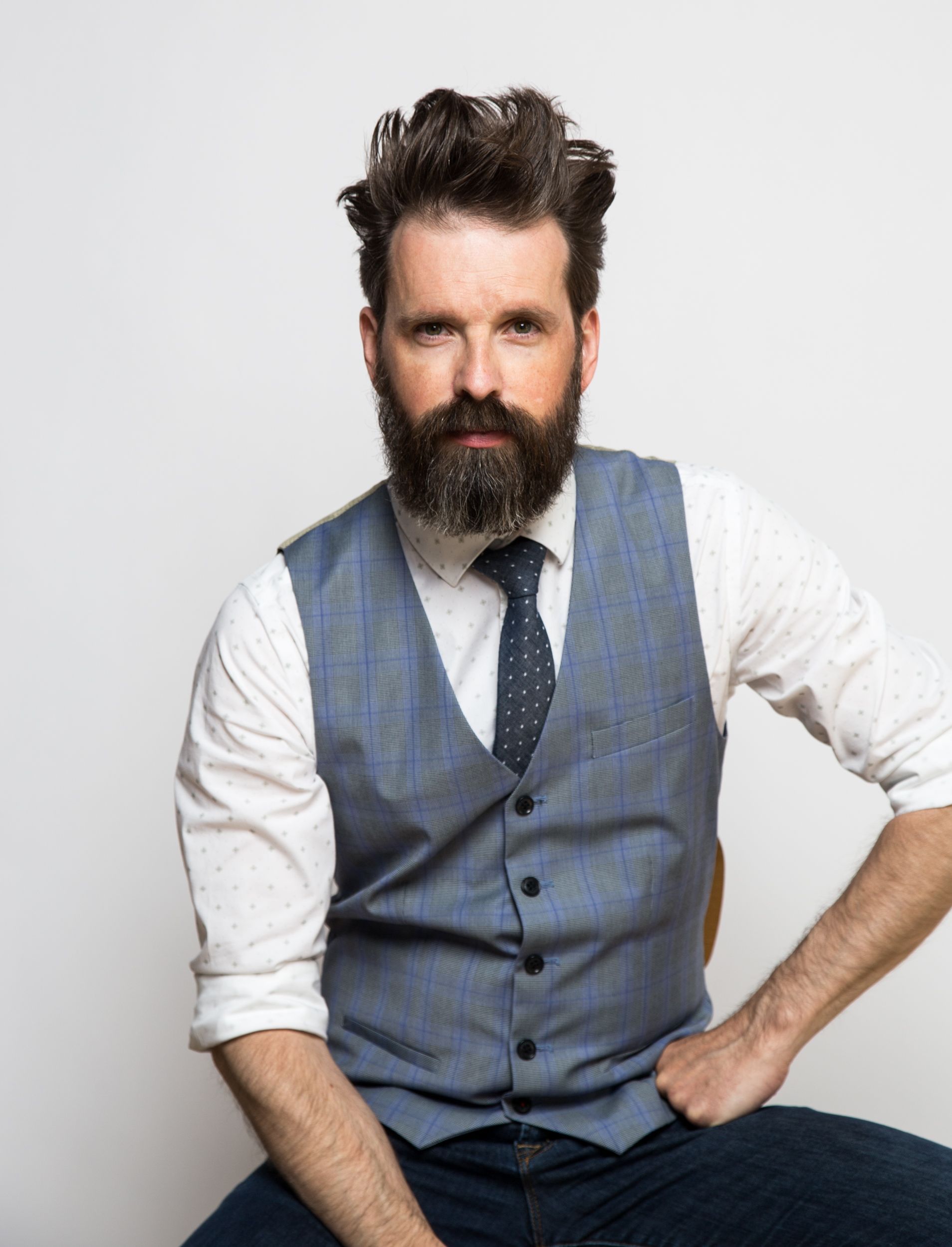 Man with beard and vest, seated, looking at camera. White shirt, blue vest and tie.