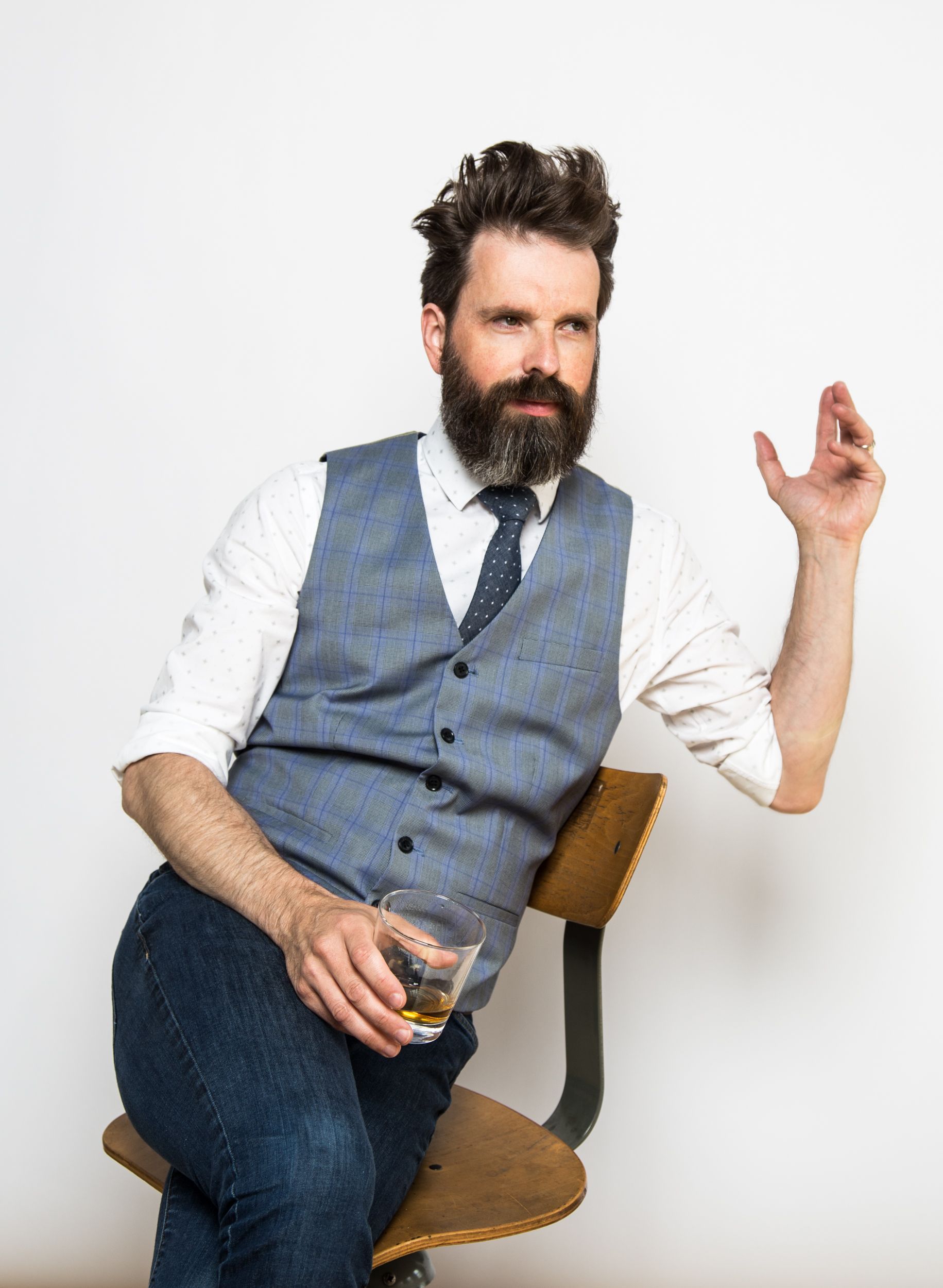 Man in vest and jeans sits, holding a drink, gesturing with his hand, looking to the side.