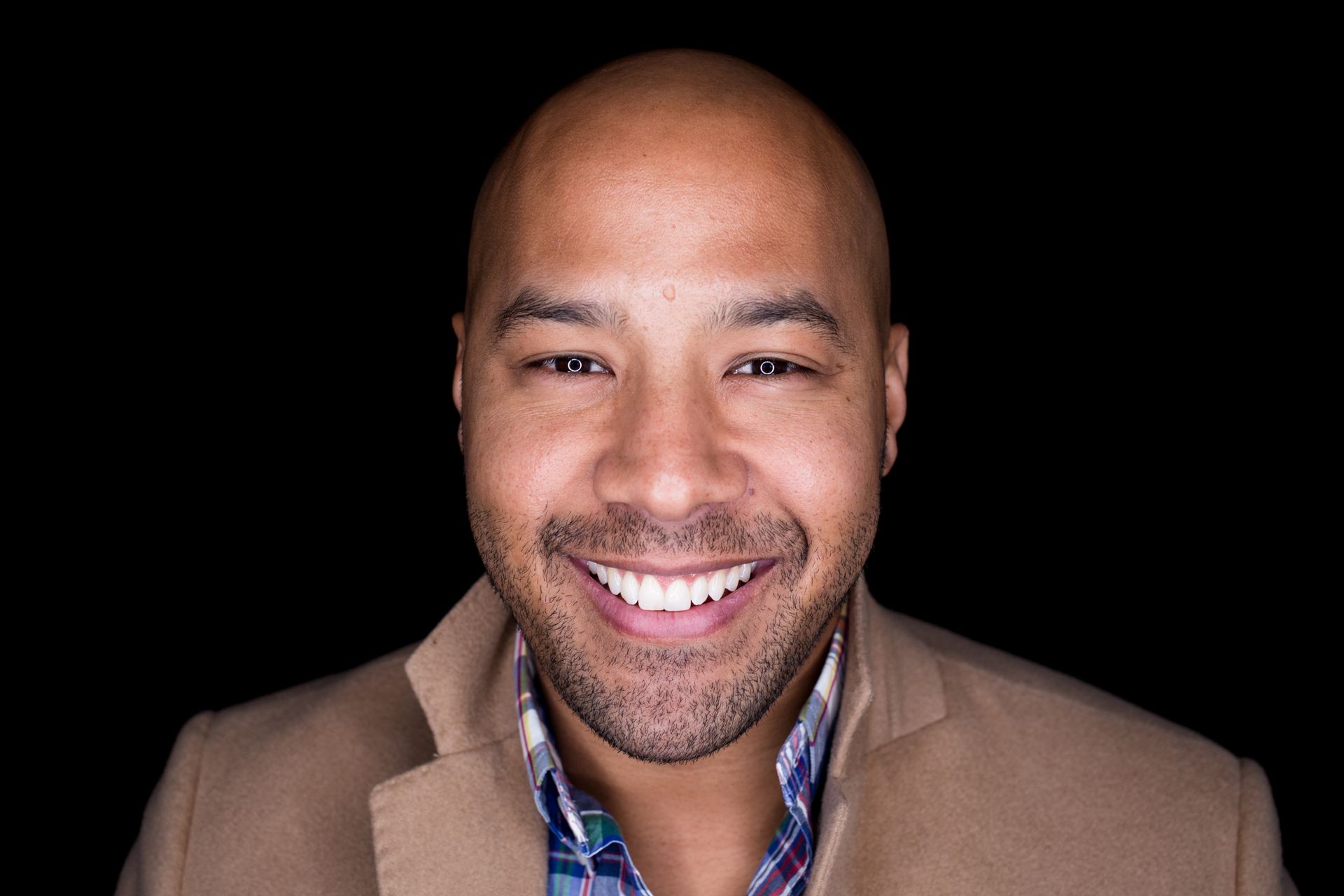 Smiling man, bald head, wearing a light brown blazer over a patterned shirt, against a black background.