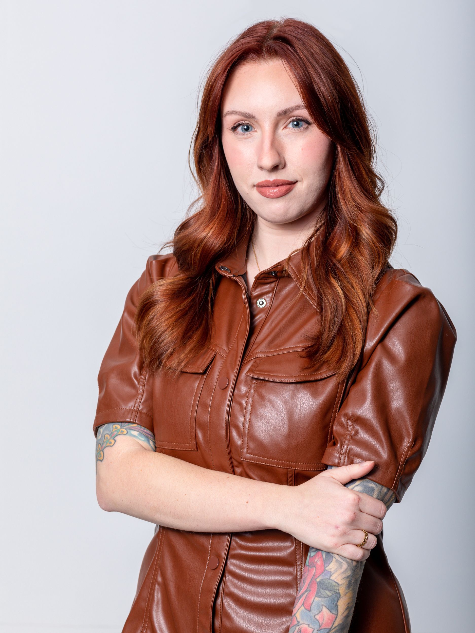 Woman with red hair, wearing a brown leather dress, arms crossed, tattoos on left arm, studio background.