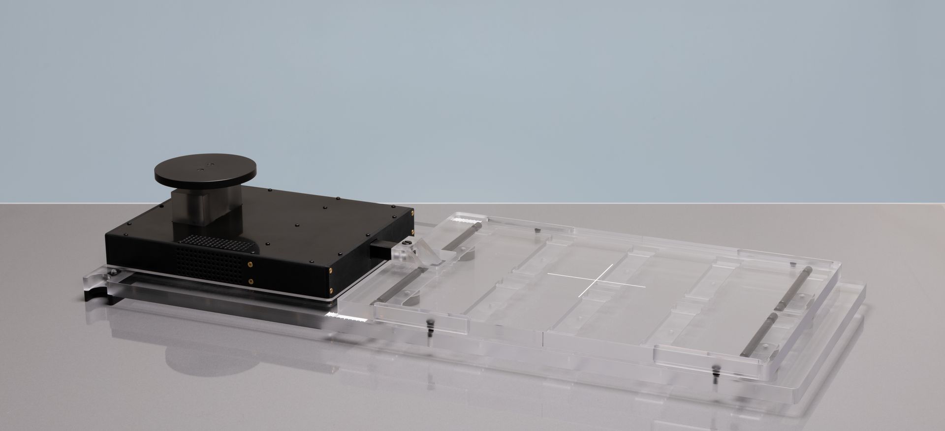 A clear plastic and black apparatus on a white surface, possibly a laboratory tool, against a blue backdrop.