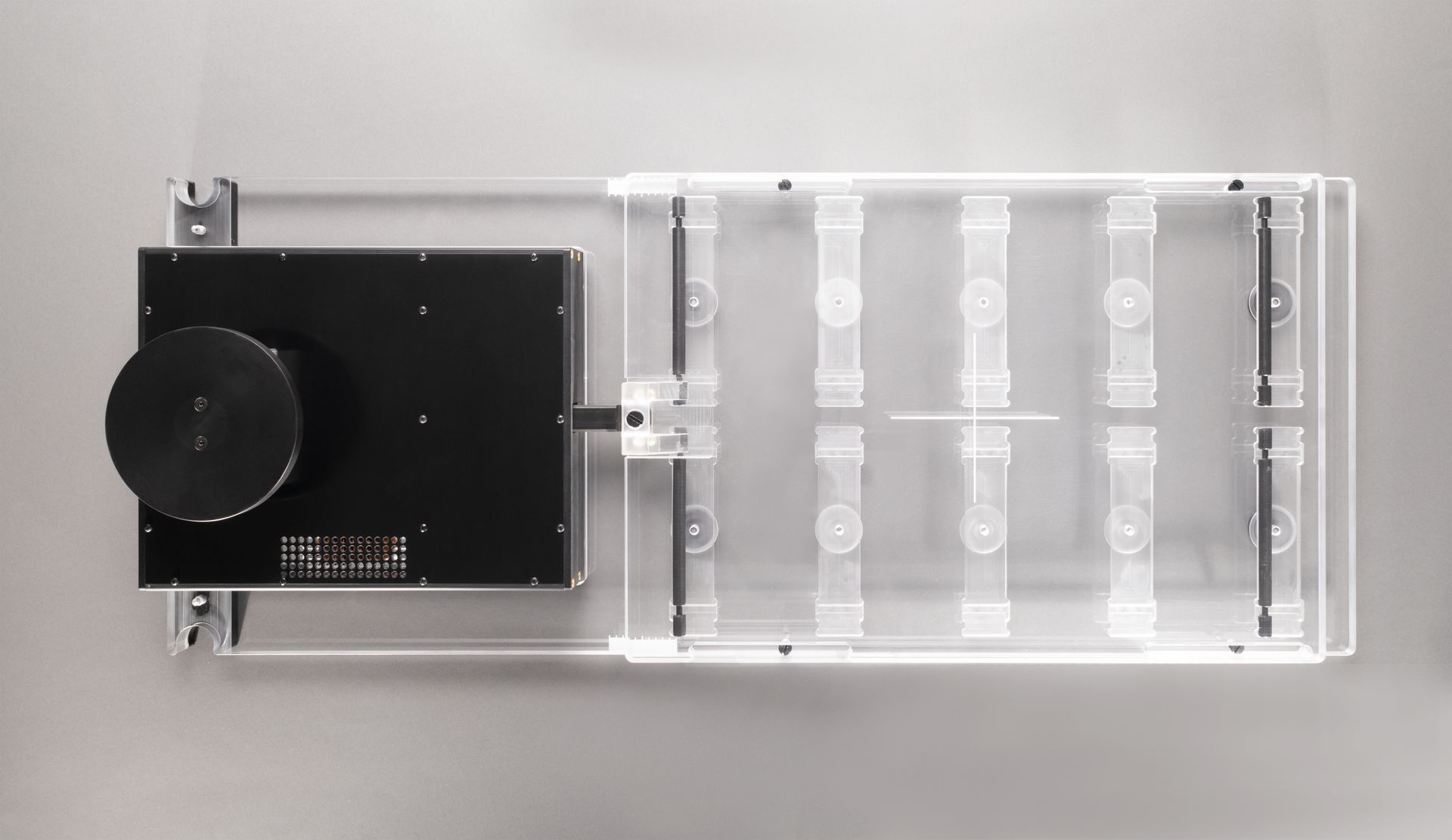 Top-down view of a rectangular, transparent enclosure with black equipment attached to the left. Mounted on a gray wall.