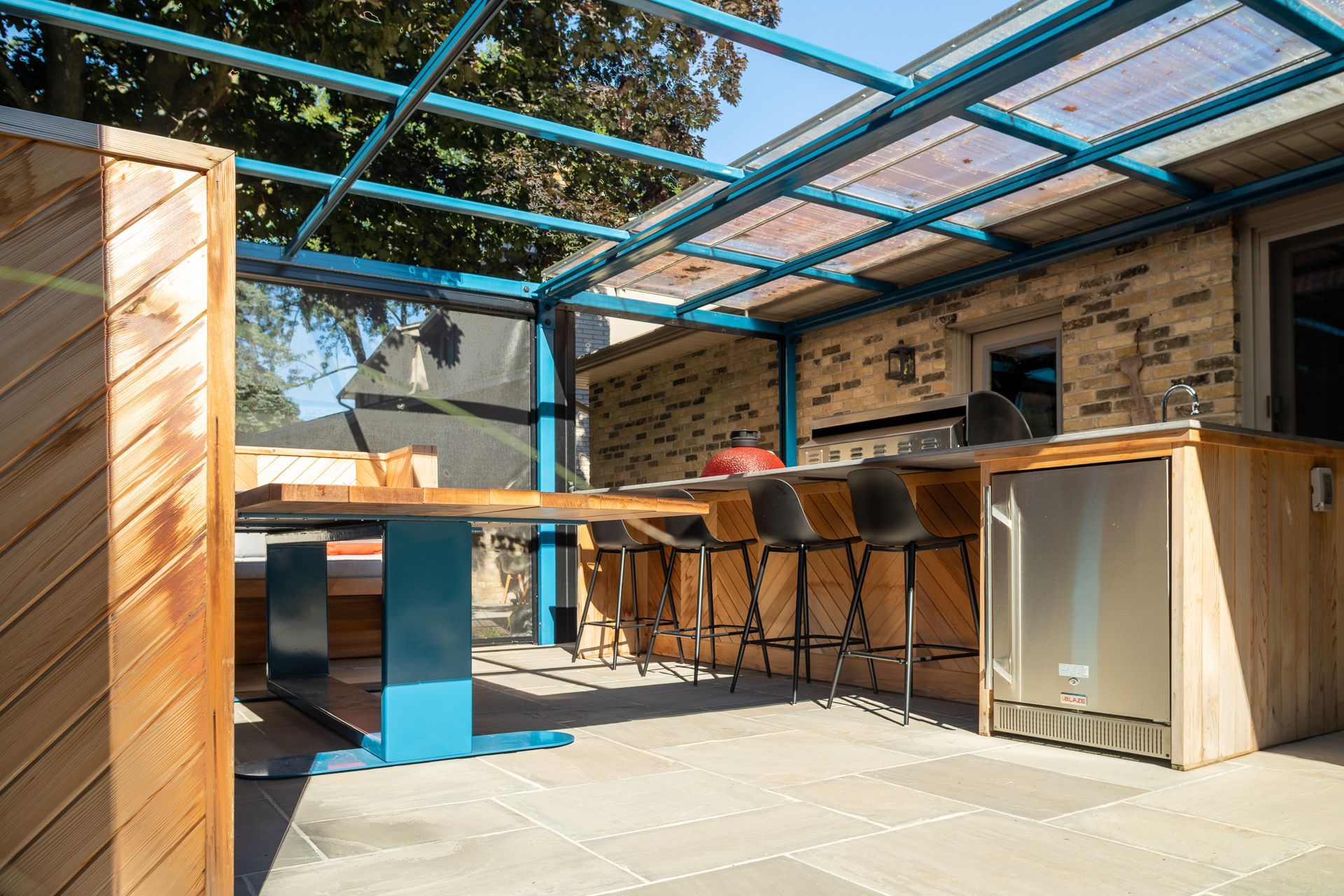 Outdoor patio with blue metal frame, bar, and table. Features stone pavers, wooden accents, and a refrigerator.