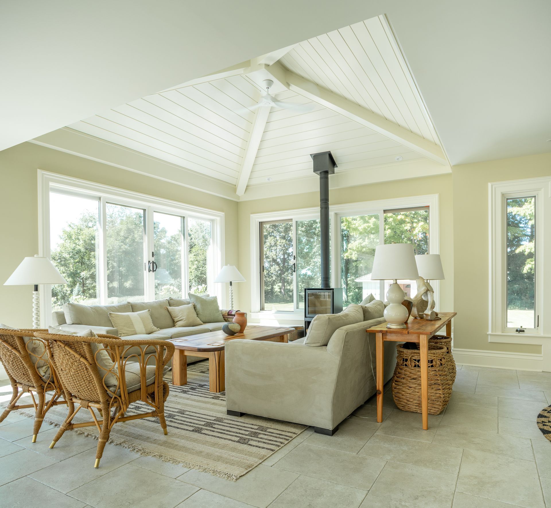 Bright sunroom with wood-burning stove, sofas, and wicker chairs, surrounded by windows with outdoor views.