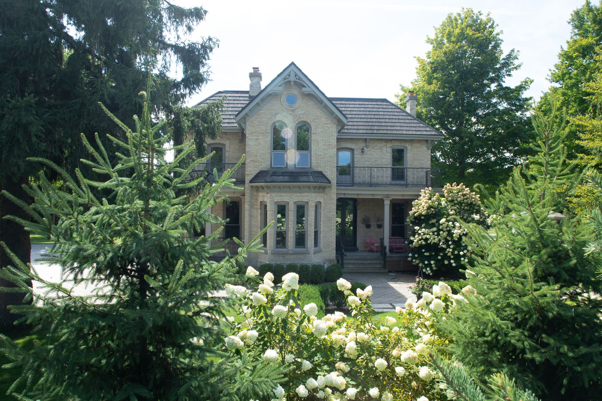 Two-story stone house with a gabled roof, surrounded by trees and shrubs in a sunny setting.