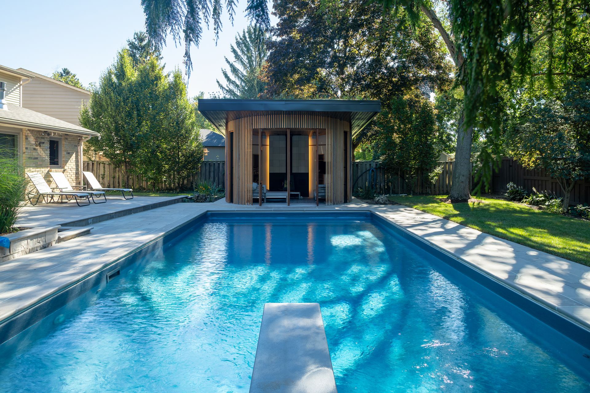 Pool with diving board, small wood-paneled cabana, and house in sunny backyard.