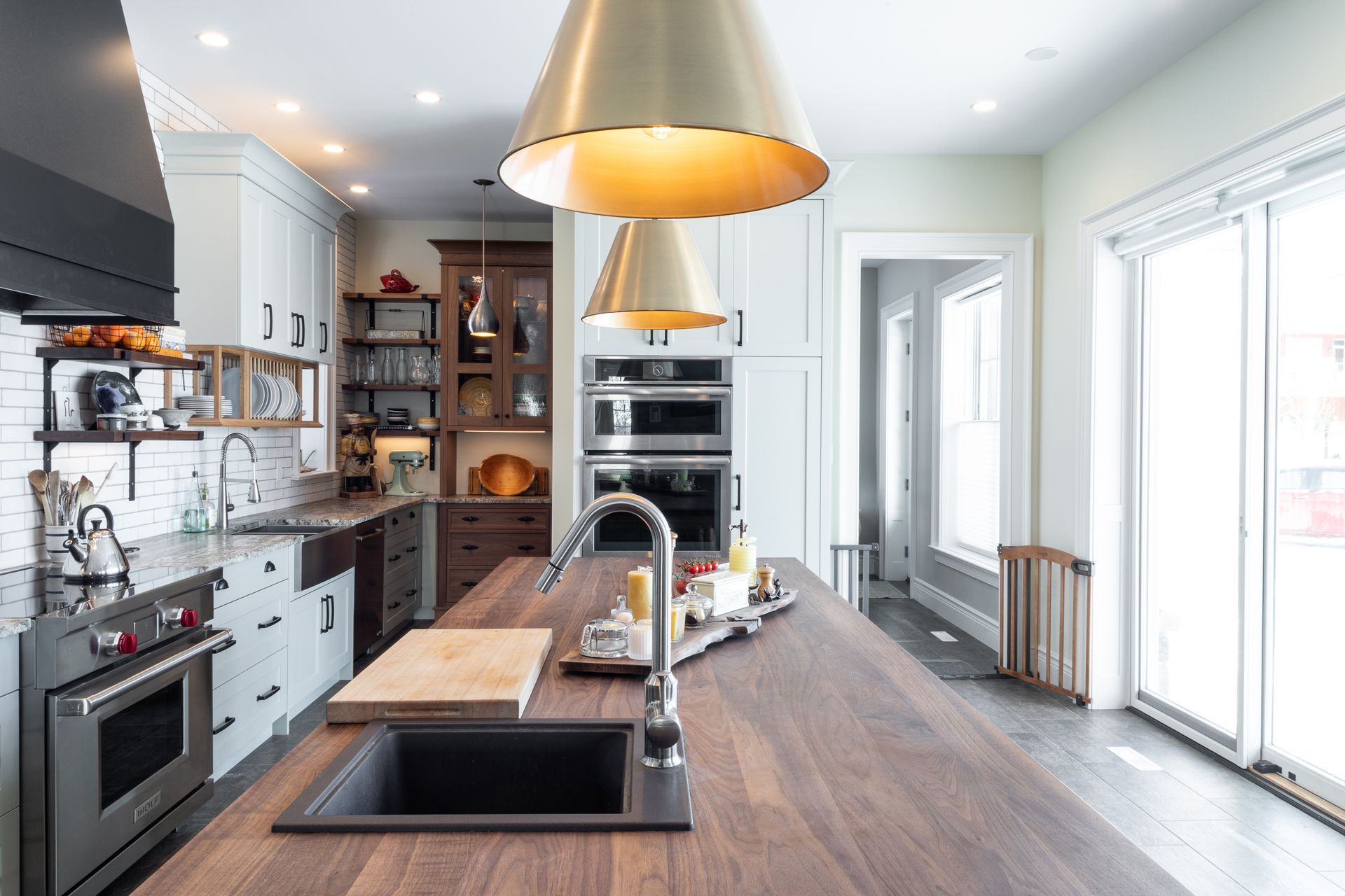 Modern kitchen with dark wood island, brass pendant lights, stainless steel appliances, and large windows.