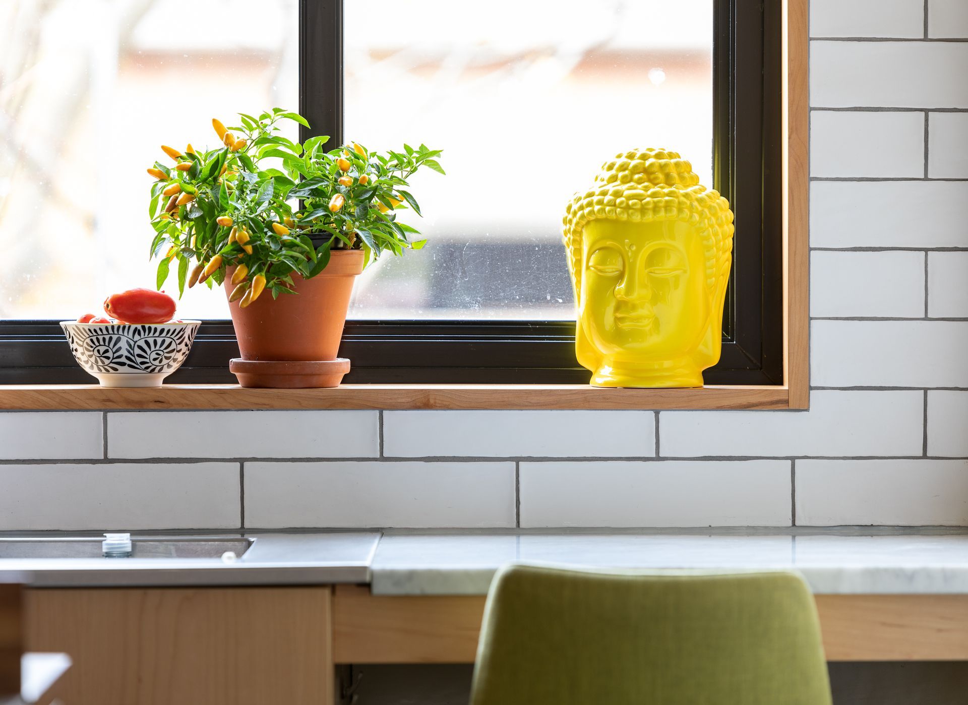 Yellow Buddha head and potted plant on a windowsill with white tile backsplash and a green chair.