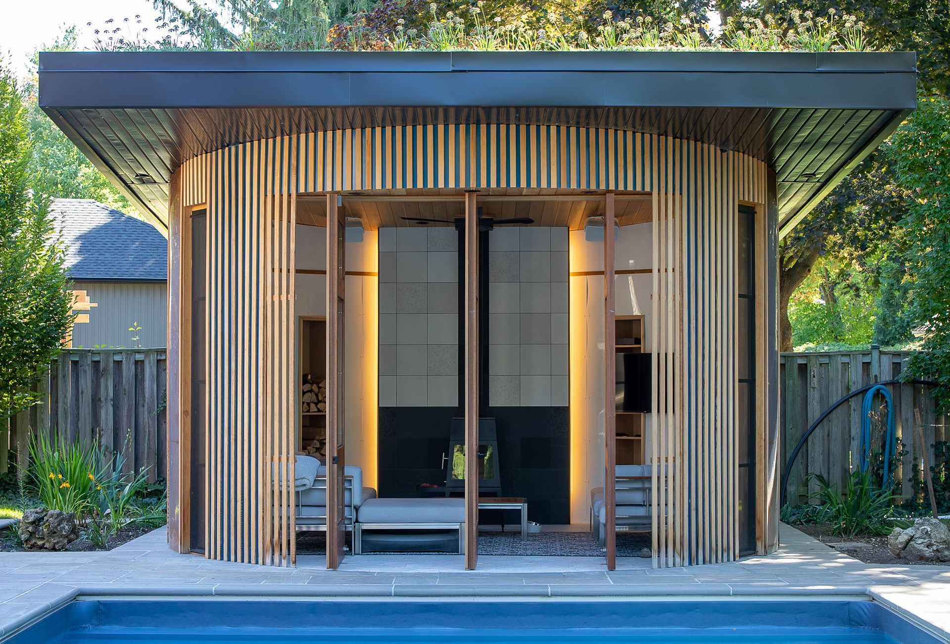 A modern, curved wooden pool house with vertical slats, open glass doors, and a flat roof, overlooking a pool.
