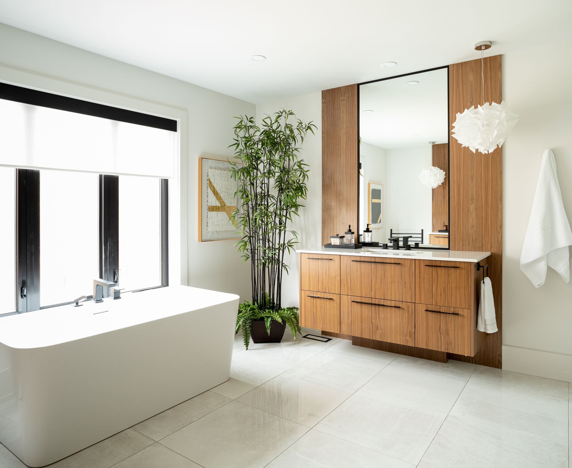 Modern bathroom with white tub, wood vanity, large mirror, and bamboo plant.