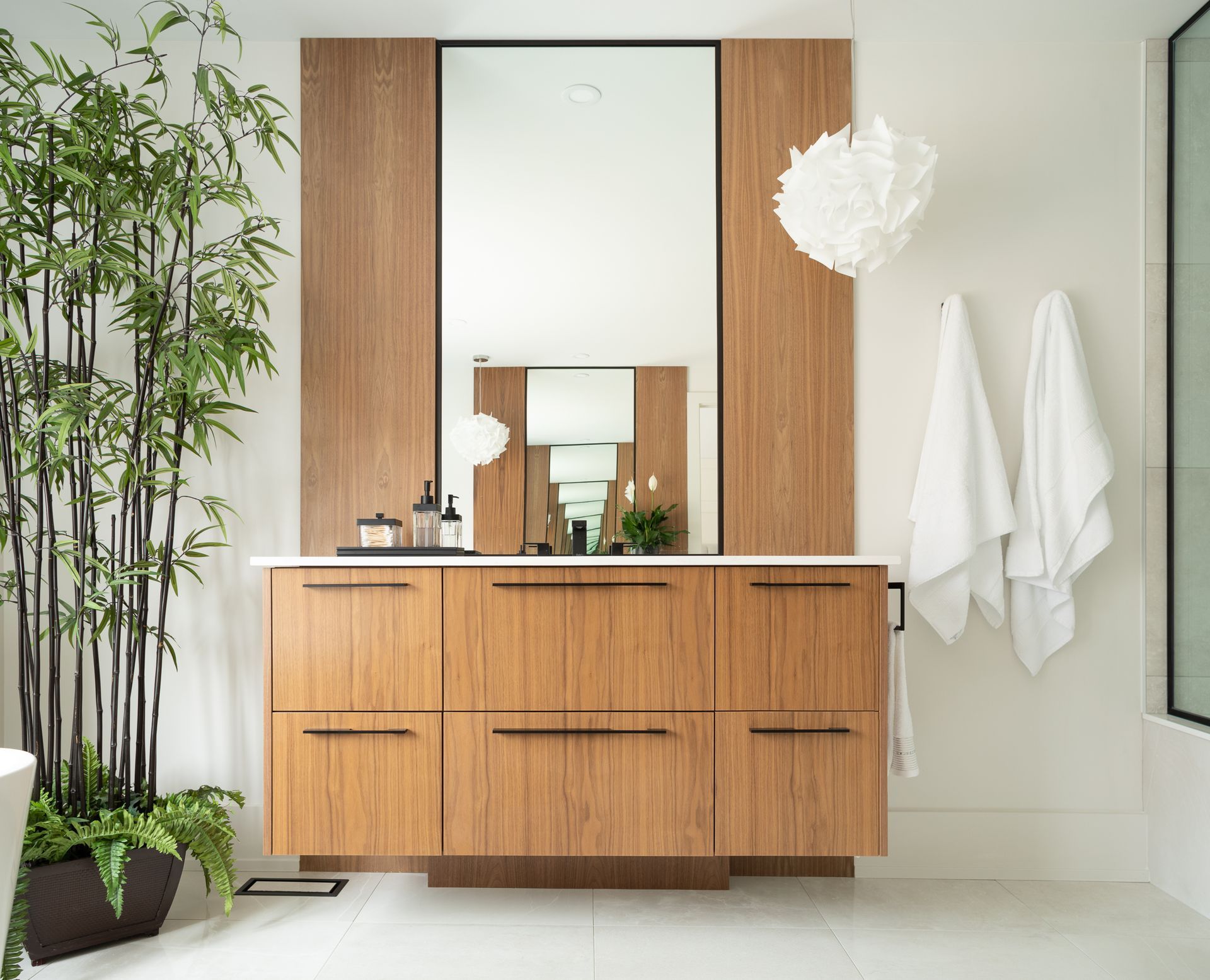 Modern bathroom with wood vanity, large mirror, white walls, and bamboo plant.