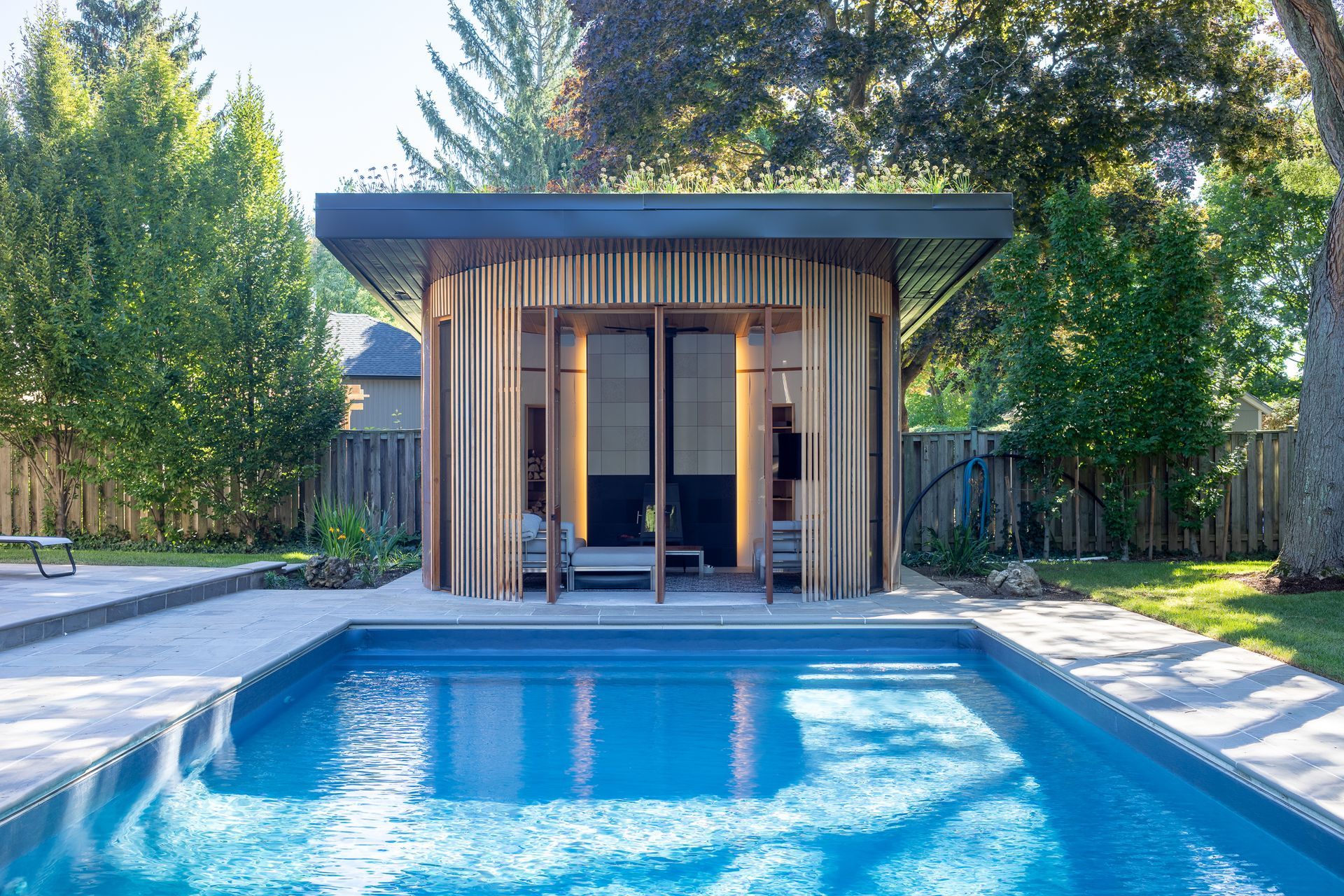 Poolside cabana with curved metallic walls, open front, and dark doors, beside a rectangular pool.