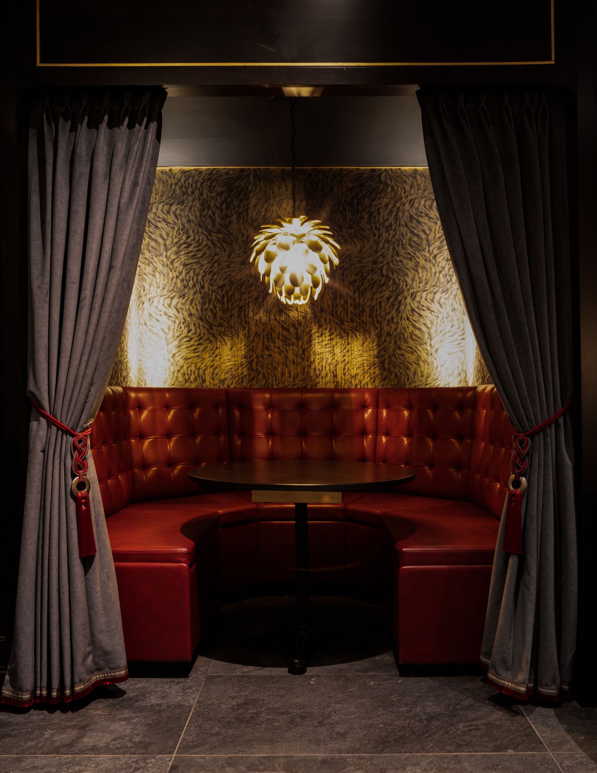Red booth with a round table, flanked by gray curtains. A gold textured wall and a decorative light fixture complete the scene.
