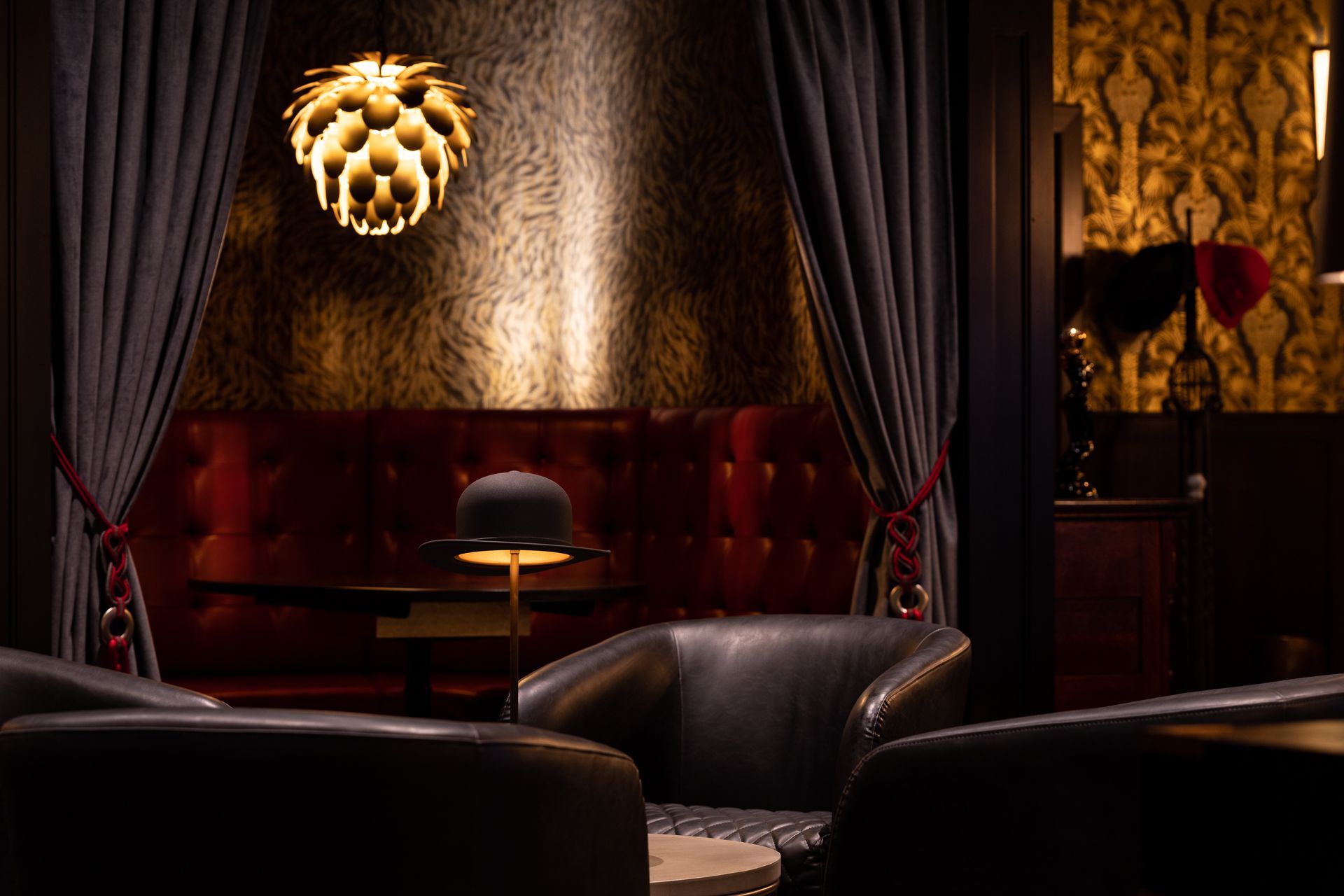 Cozy, dark-lit lounge with velvet booth, leather armchairs, gold patterned wallpaper, and a unique pendant lamp.