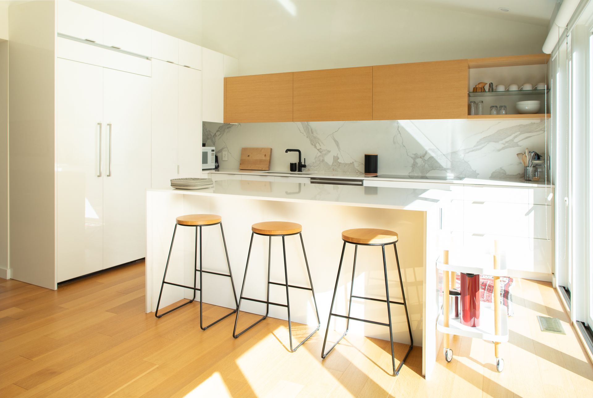 Modern kitchen with a breakfast bar, stools, and built-in white cabinets. Sunny with wood flooring.