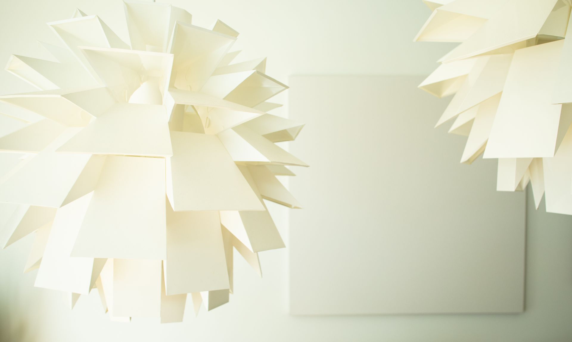 Two spiky, paper-like lamps flank a blank, square canvas on a light-colored wall.