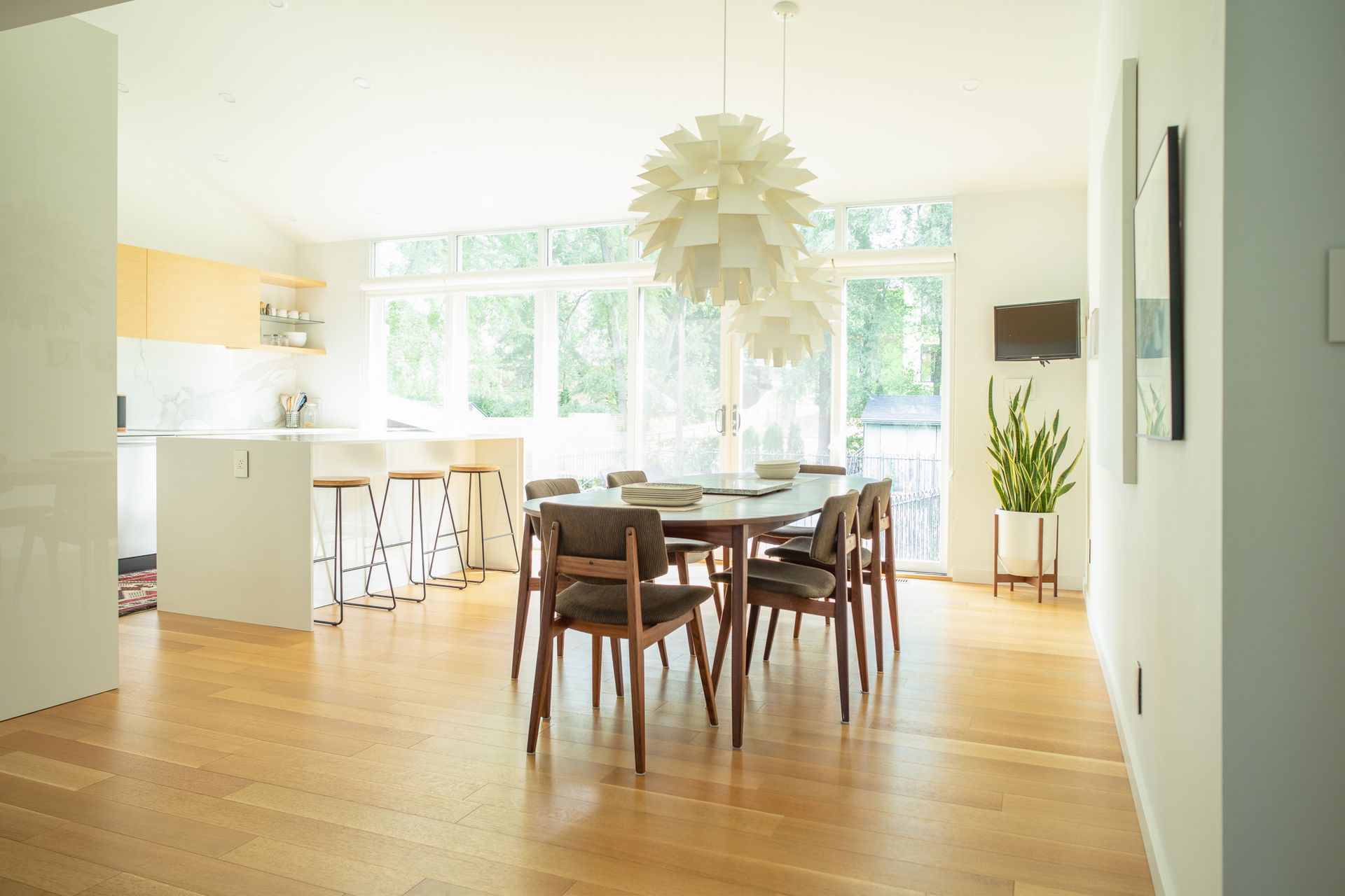 Dining room with wooden floor, table, chairs, and large white pendant light. Kitchen is visible in the background.