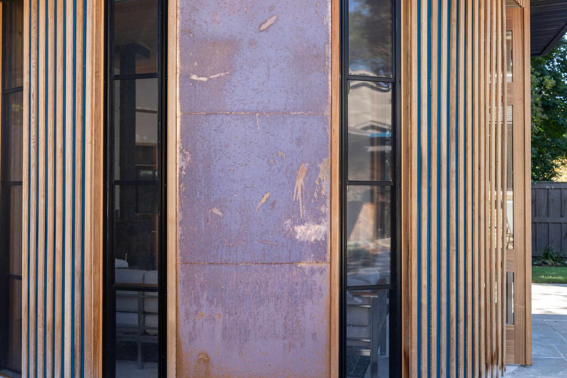 Exterior of a building with weathered copper panels and vertical wooden slats. Black-framed windows reflect the outdoors.