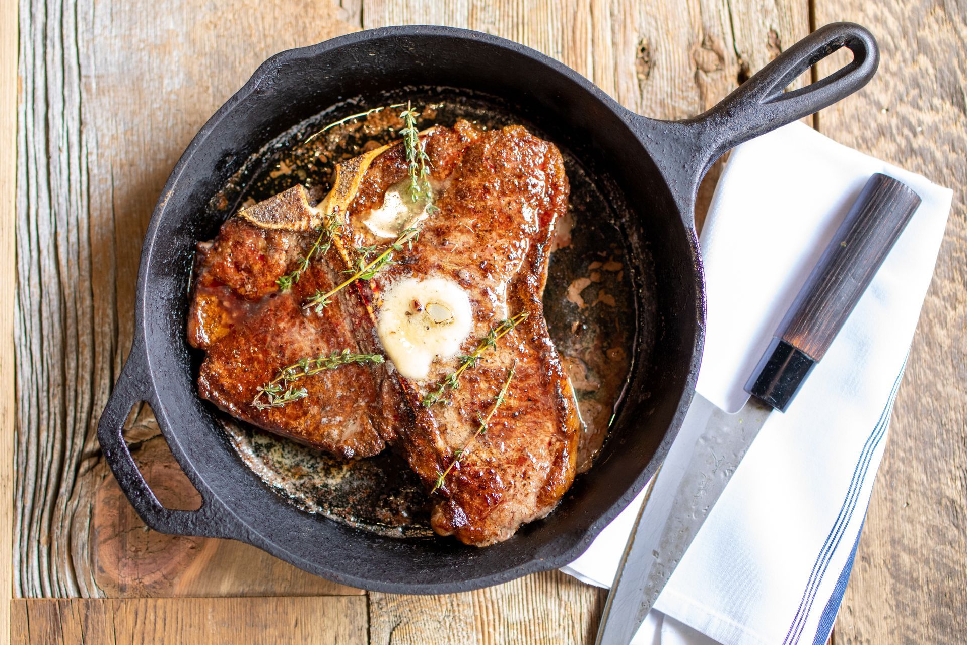 Steak in a cast iron skillet with butter and herbs on wooden surface, knife and white cloth nearby.