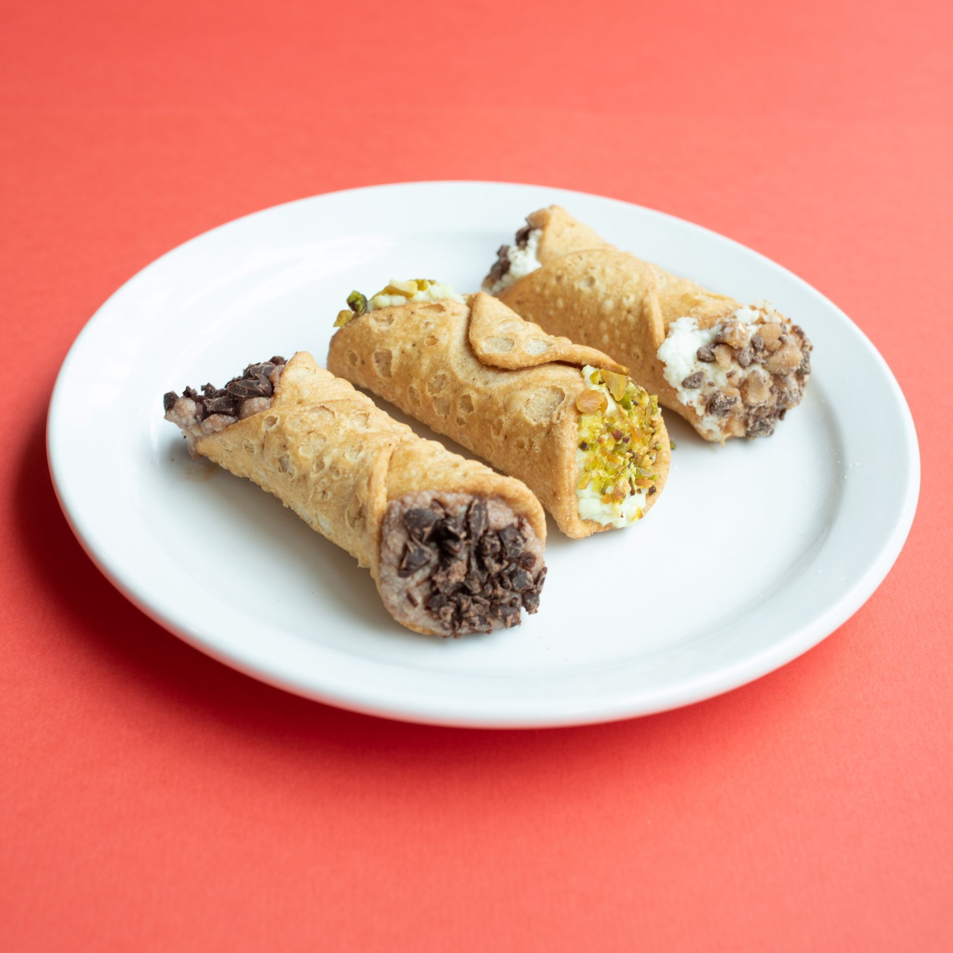 Three cannolis on a white plate, with chocolate chips, pistachios, and nuts, on a coral background.