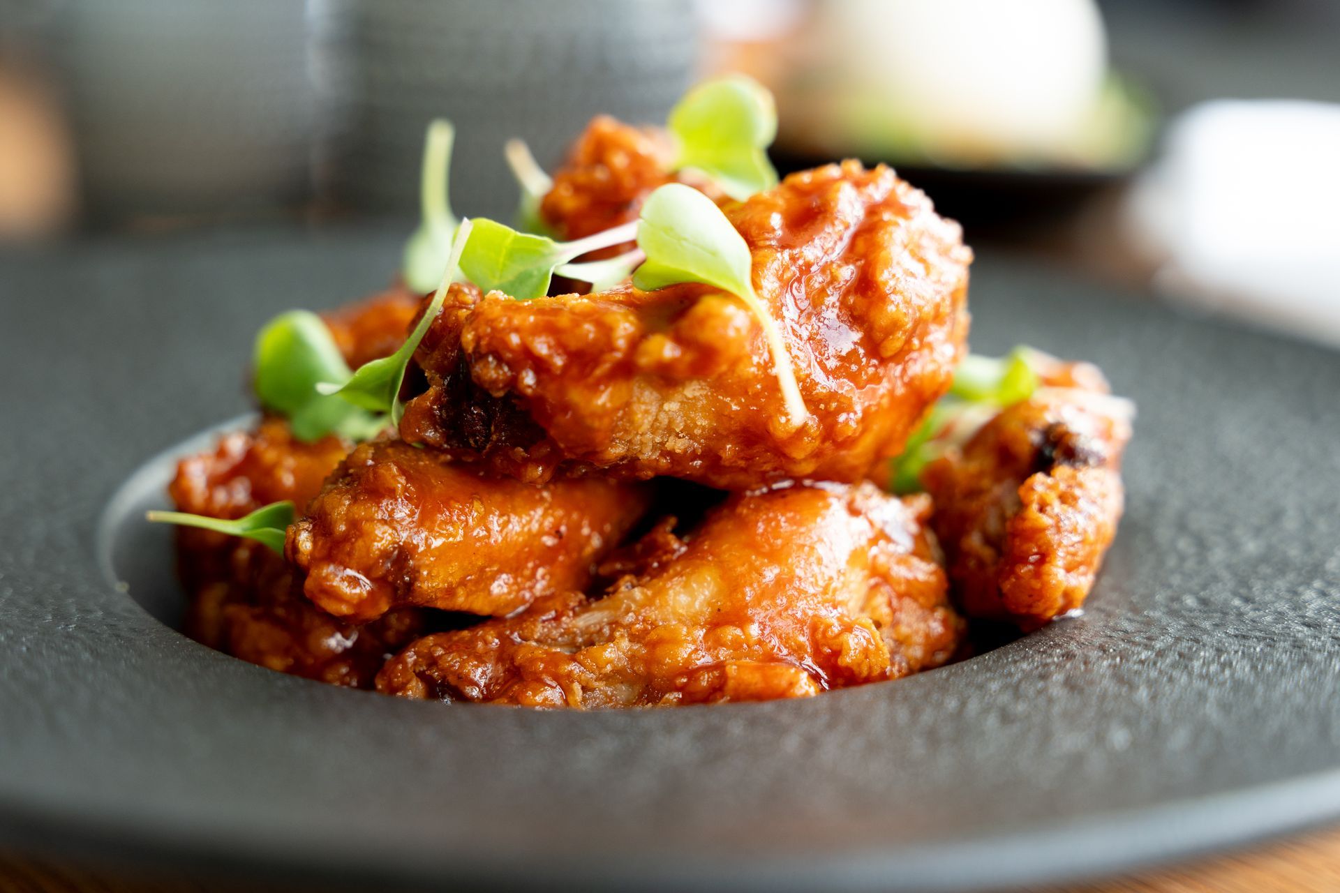 Fried chicken pieces in a red sauce, topped with sprouts, on a dark plate.